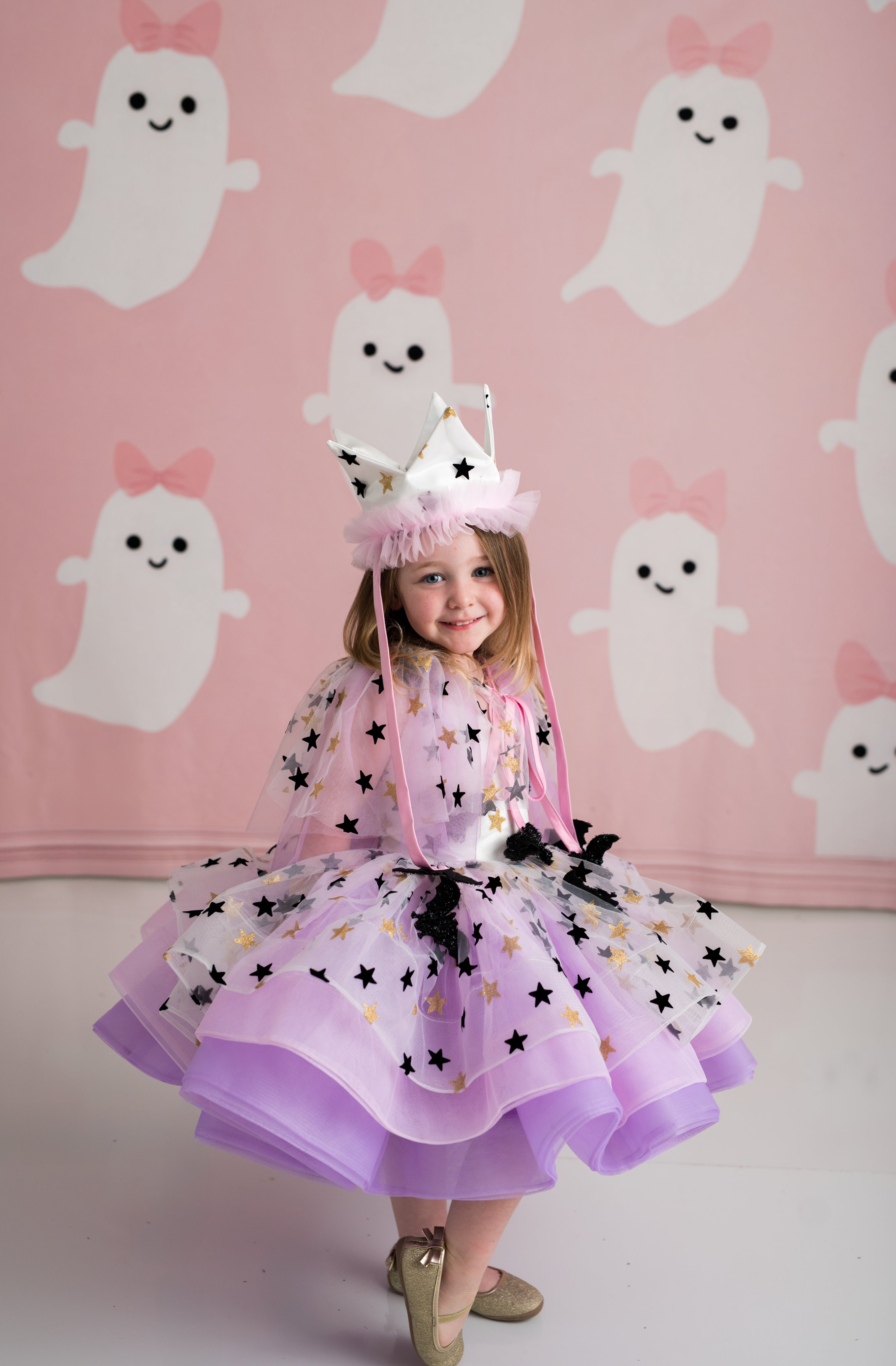 Child in a pink and purple dress with star patterns against a pink background with ghost illustrations.