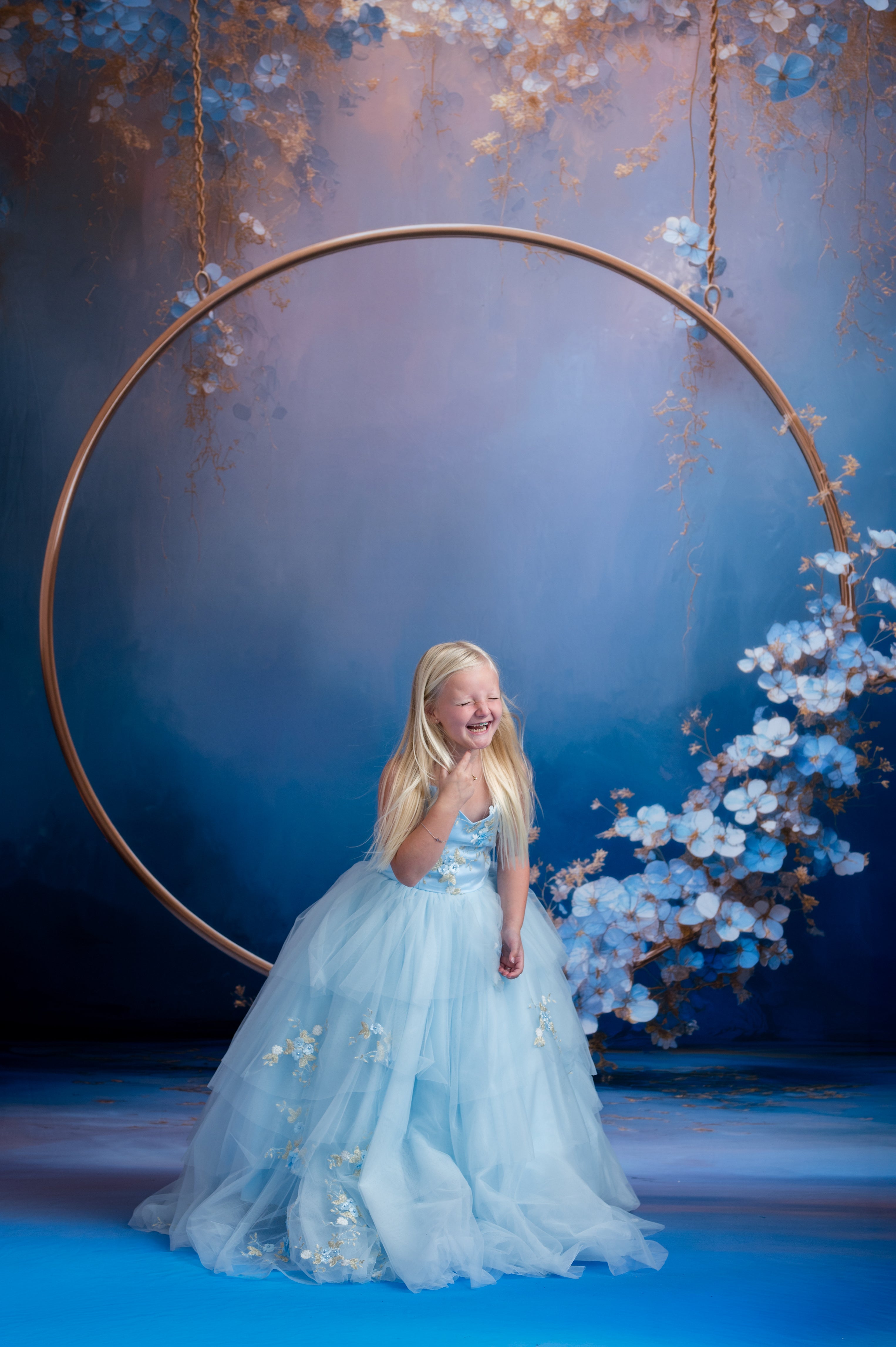 Young girl in a light blue dress standing in front of a large circular frame with floral decorations on a blue background.