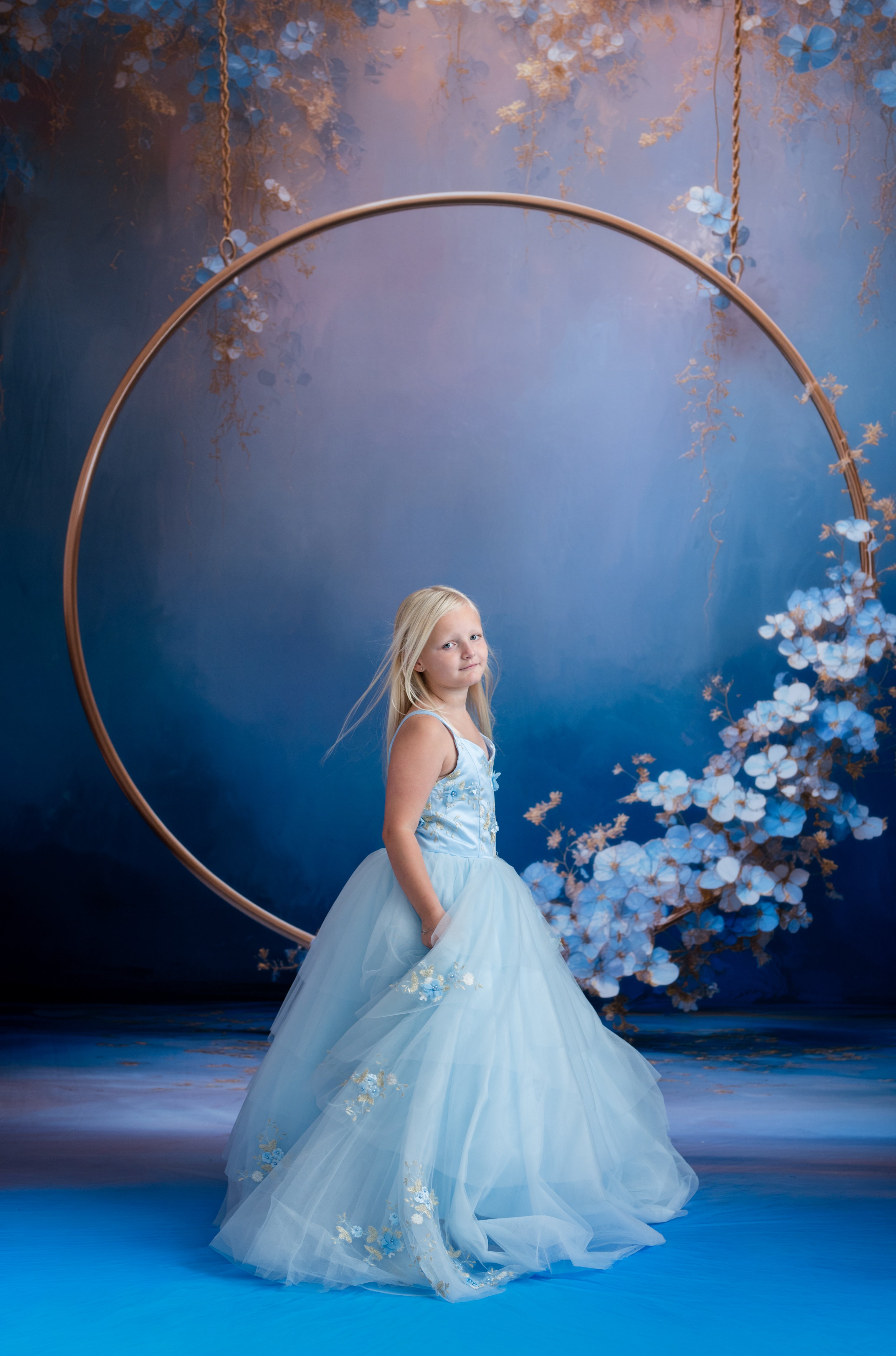 Young girl in a light blue dress standing in front of a large circular frame with floral decorations.