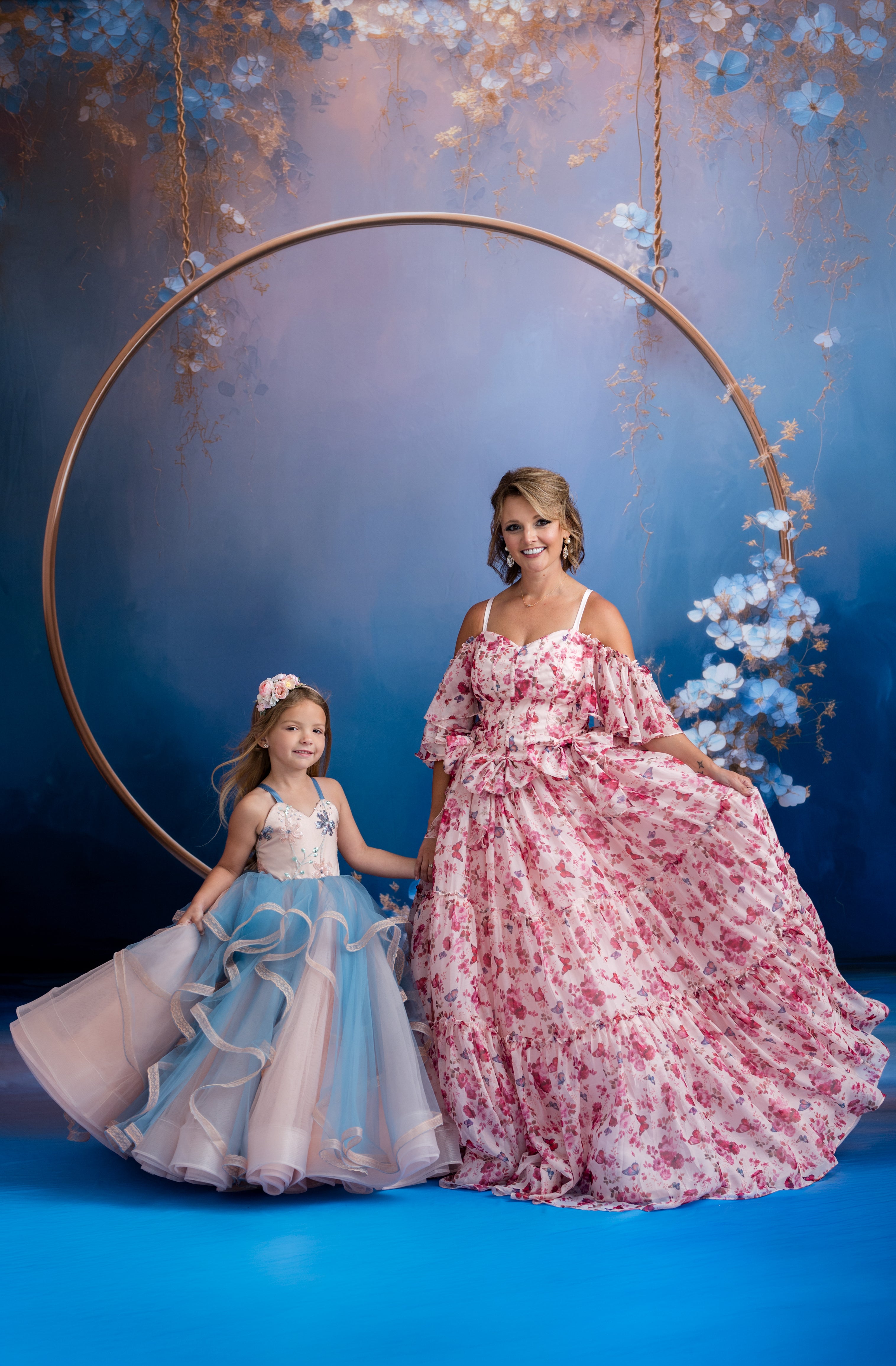 Woman and child in formal dresses standing in front of a floral hoop against a blue background