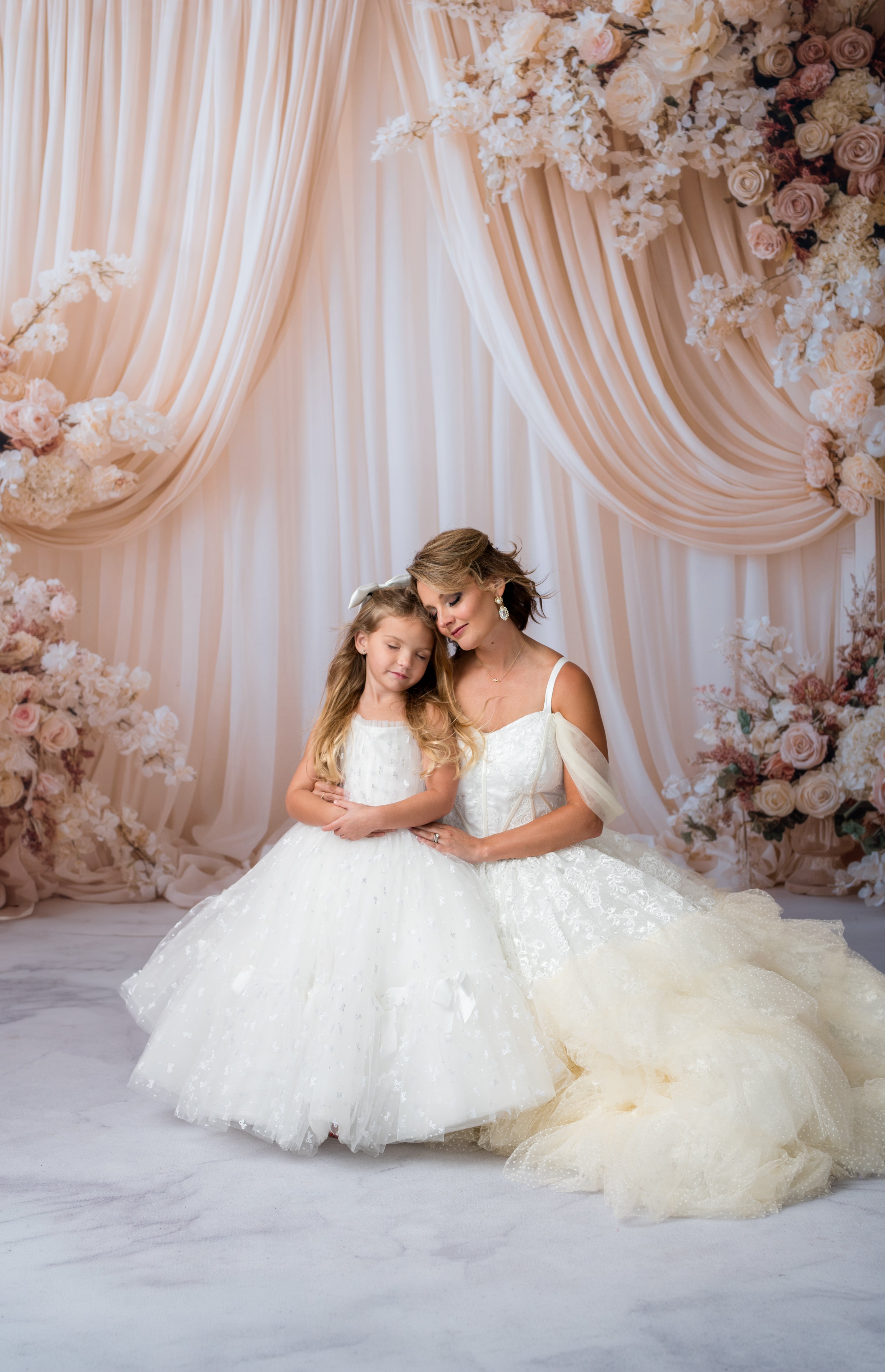 Woman and young girl in white dresses sitting together with floral decorations in the background