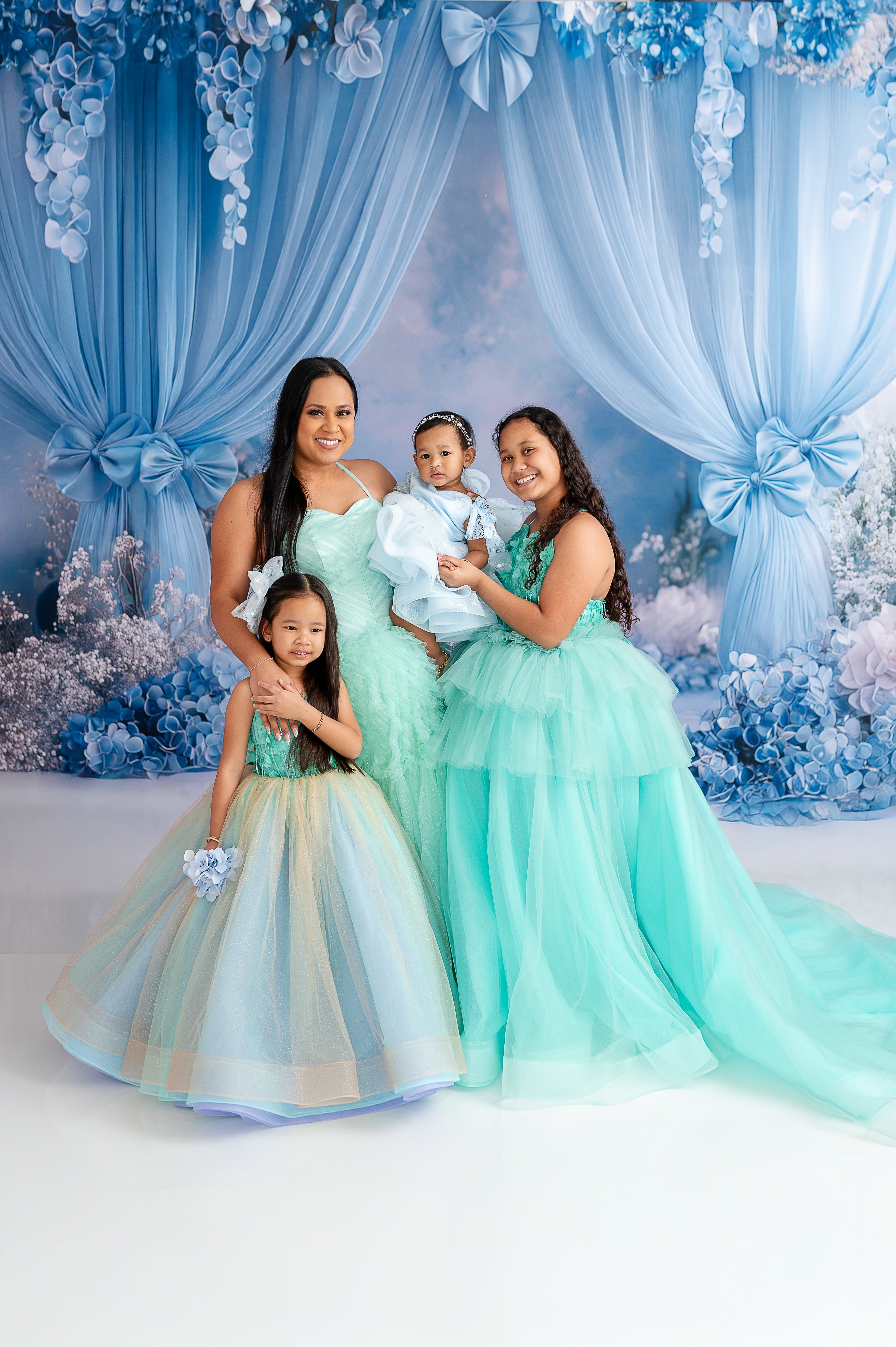 "Mother and daughter in elegant aqua and mint tulle gowns posing in front of a dreamy blue floral backdrop."