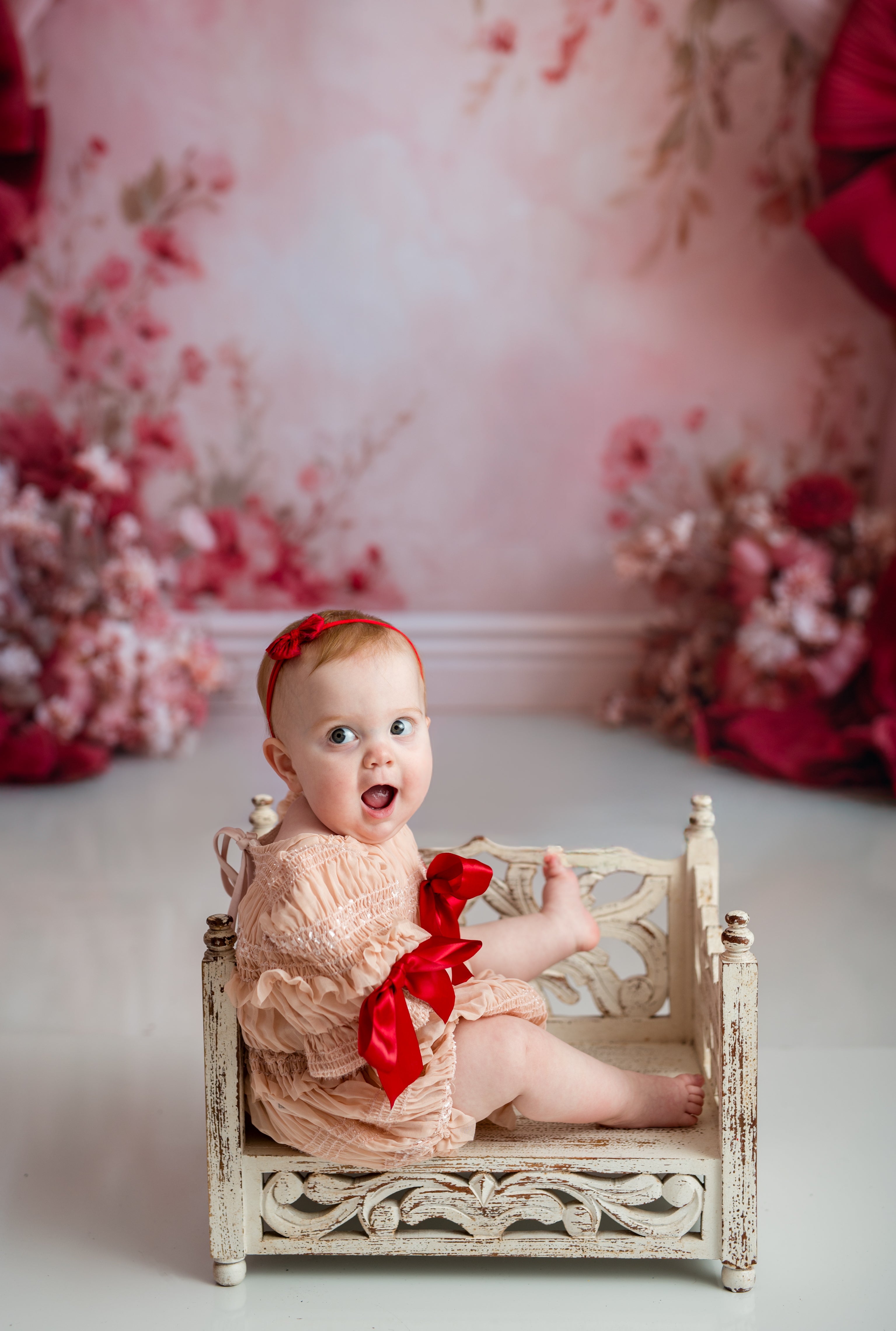 Baby sitting on a small bed with a red bow, floral backdrop