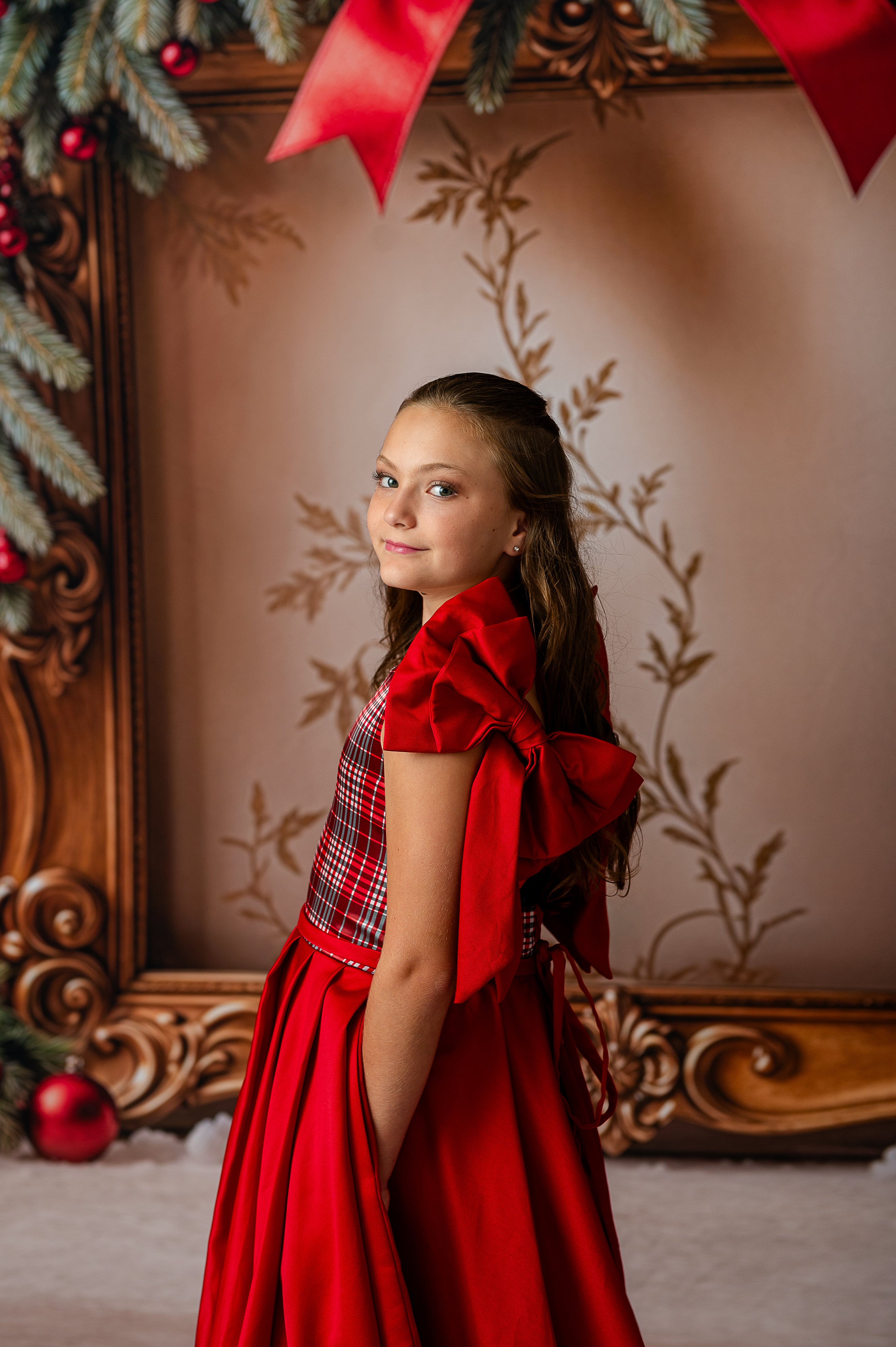 Young girl in a red dress with a large bow standing in front of a decorated Christmas tree.