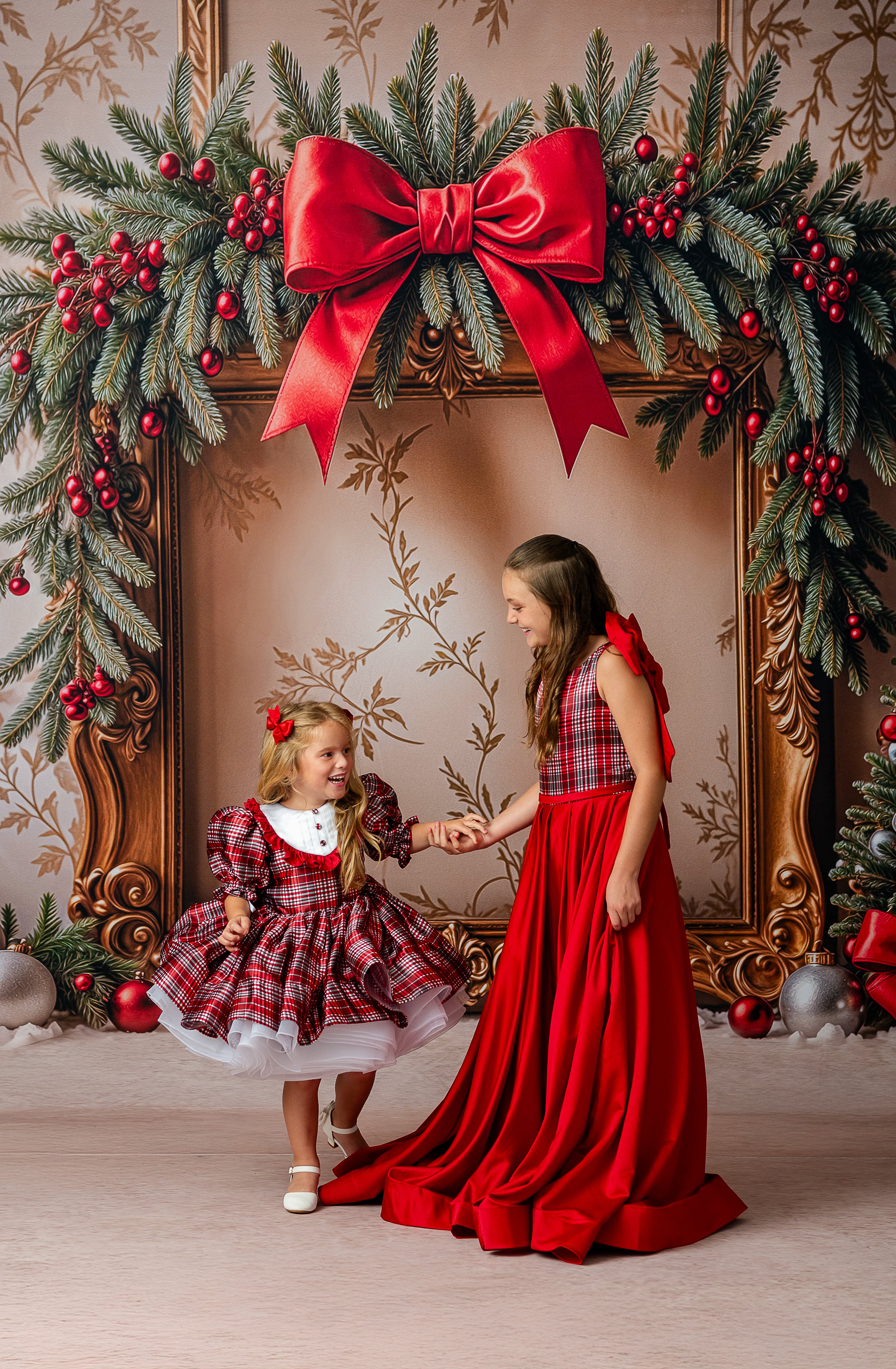 Two children in red and plaid dresses standing in front of a decorated Christmas wreath with a large red bow.