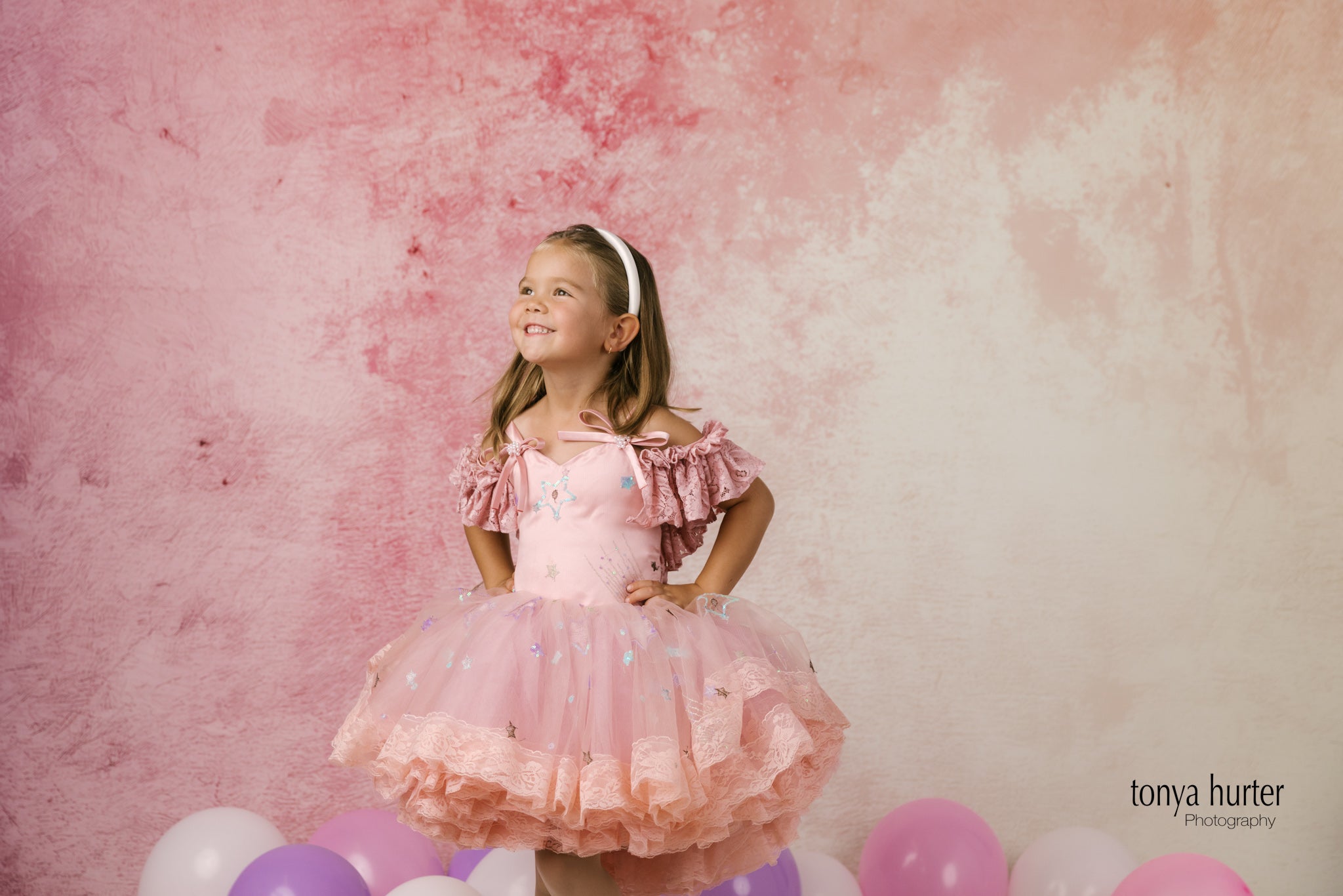 Young girl in a pink dress standing against a pink and white textured background with balloons.