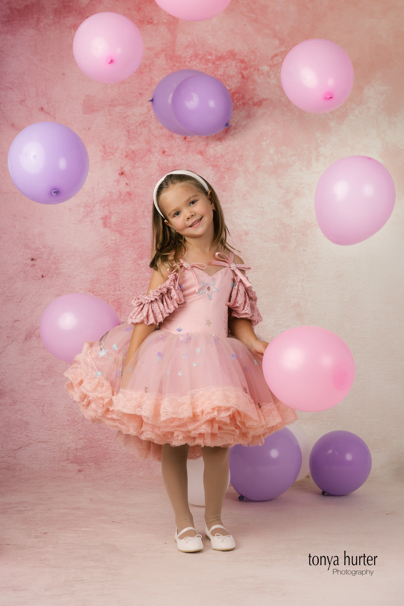 Young girl in a pink dress surrounded by pink and purple balloons against a pink background