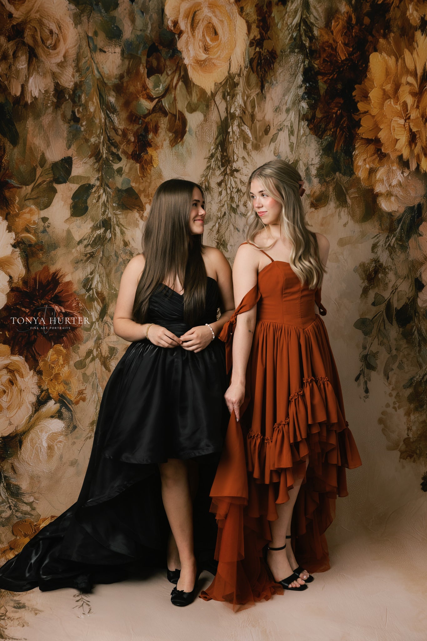 Two women in black and rust dresses standing against a floral backdrop
