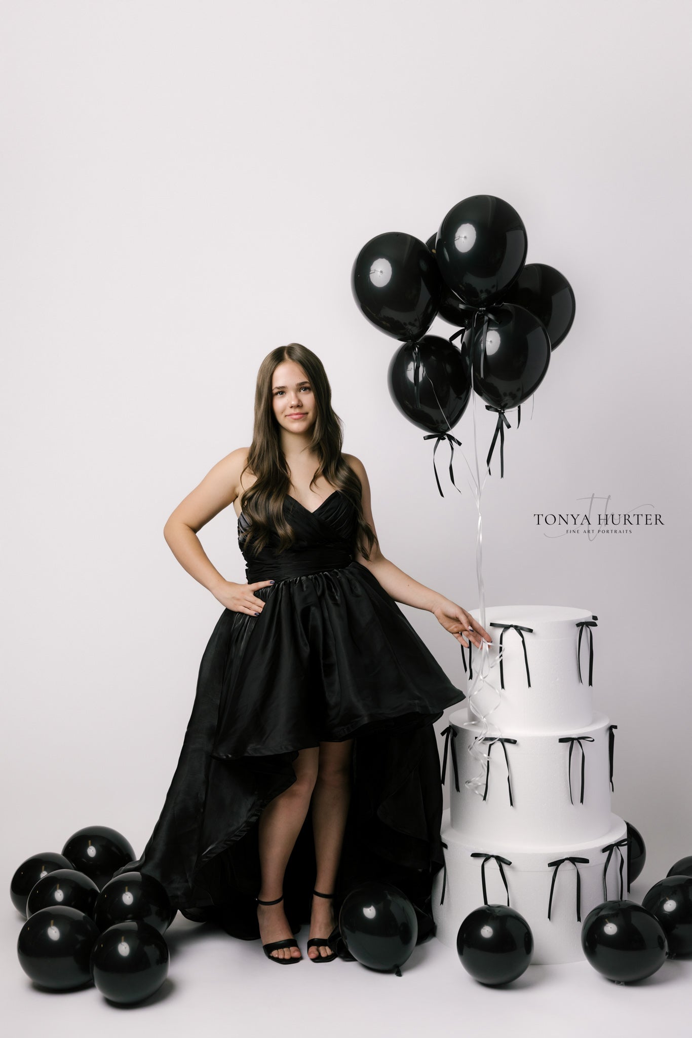 Woman in a black dress standing next to black balloons and a cake with 'TONYA HURTER' branding.