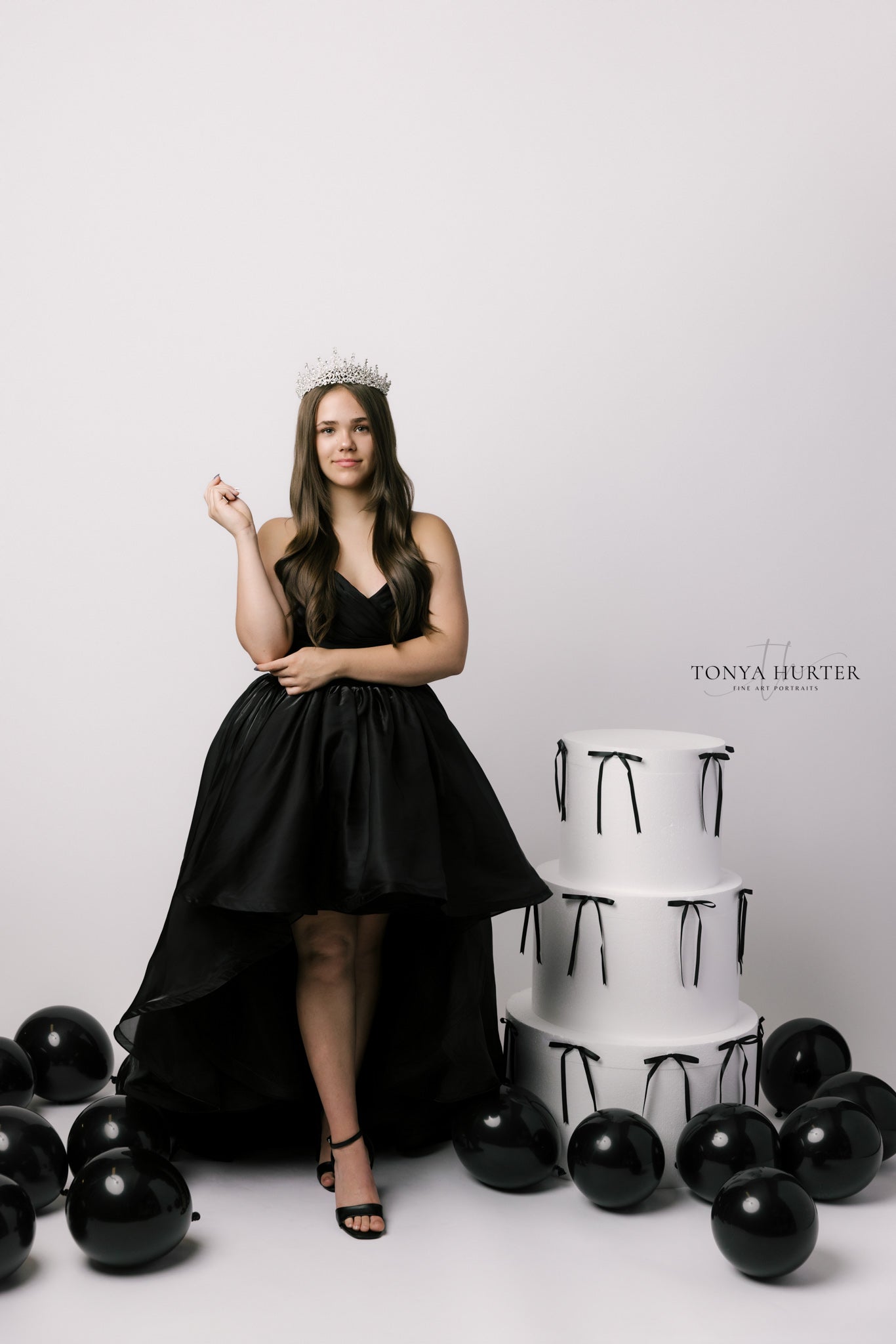 Woman in a black dress standing next to a white cake with black ribbons on a white background.
