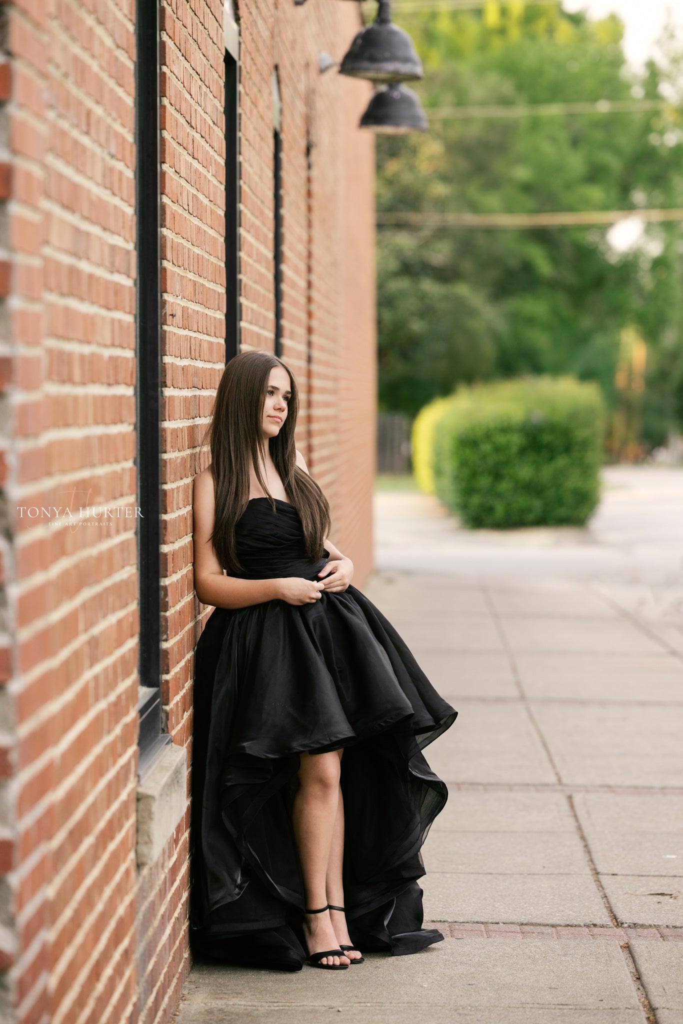 Woman in a black dress standing against a brick wall on a sidewalk.