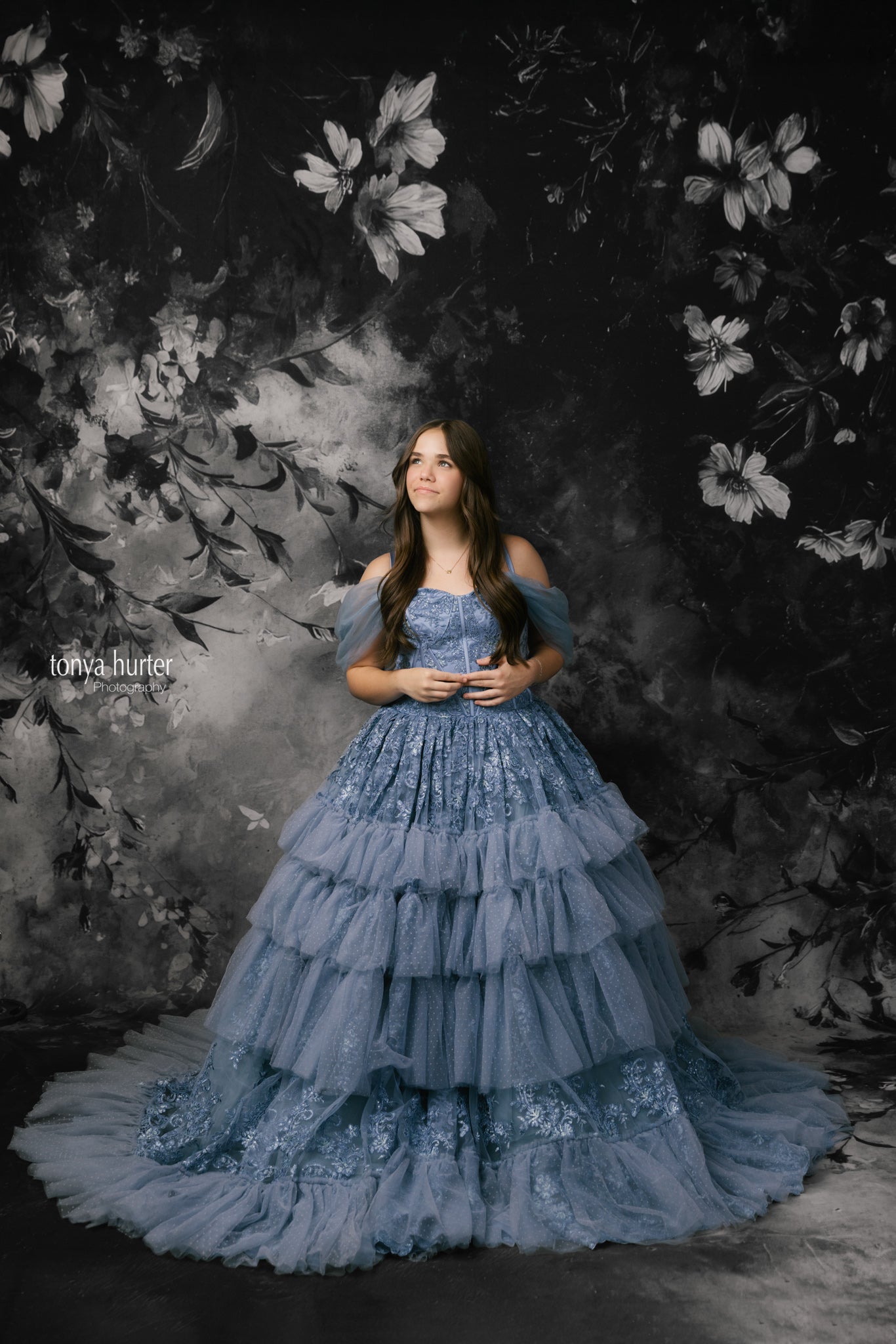 Woman in a blue gown standing against a dark floral backdrop