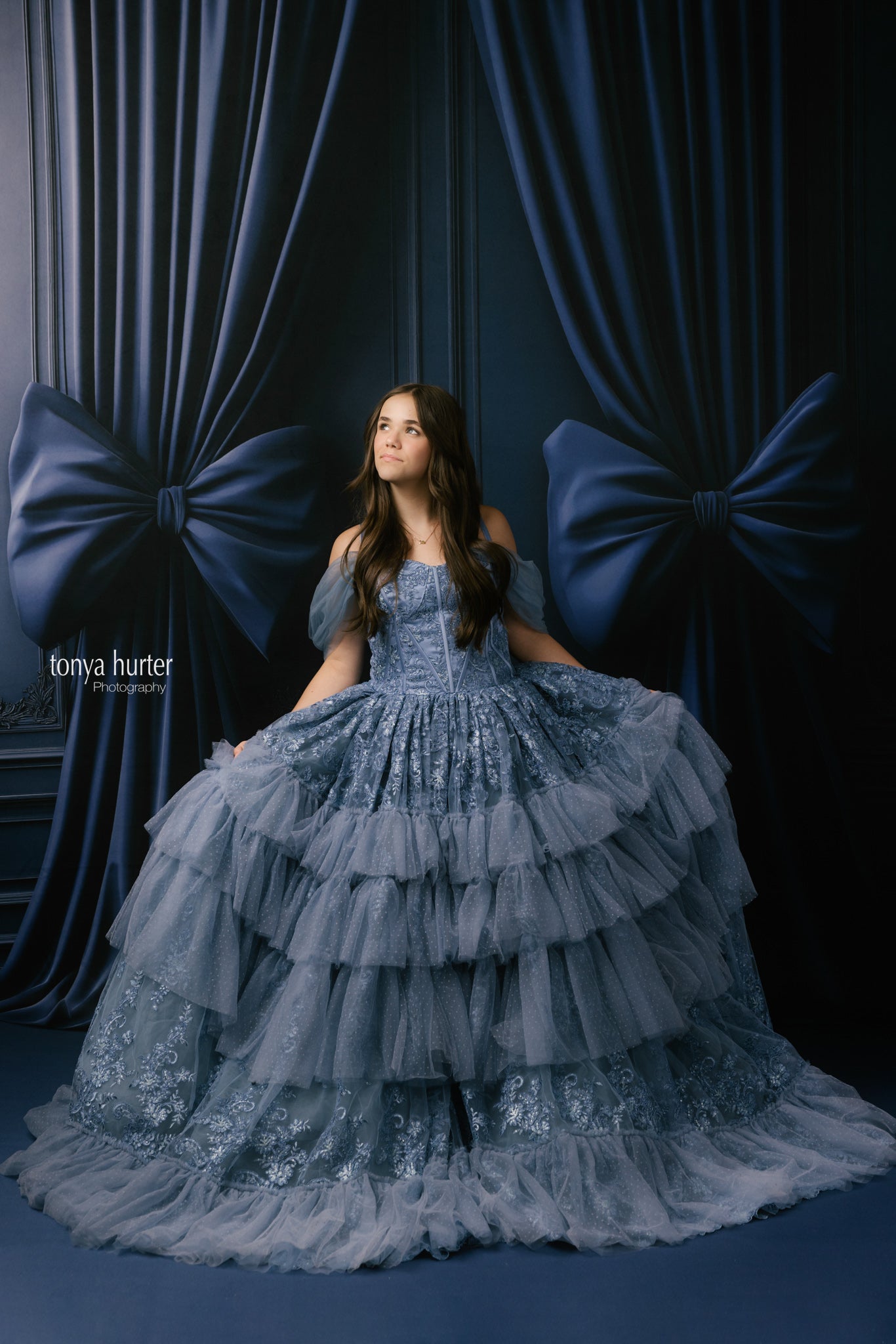 Woman in a blue tiered gown standing against a dark curtain backdrop