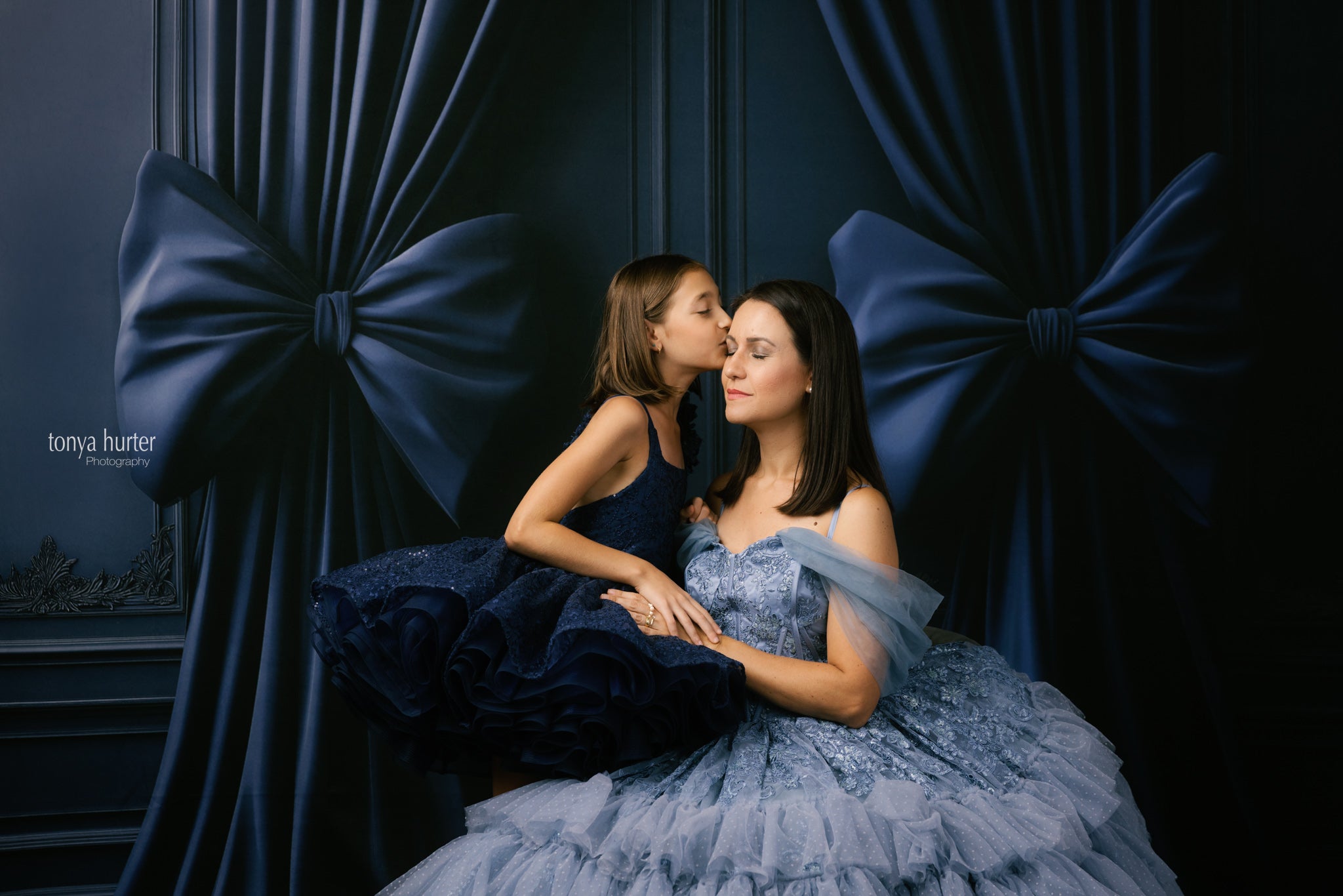 Two women in formal dresses sitting close together against a dark blue curtain with large bows.