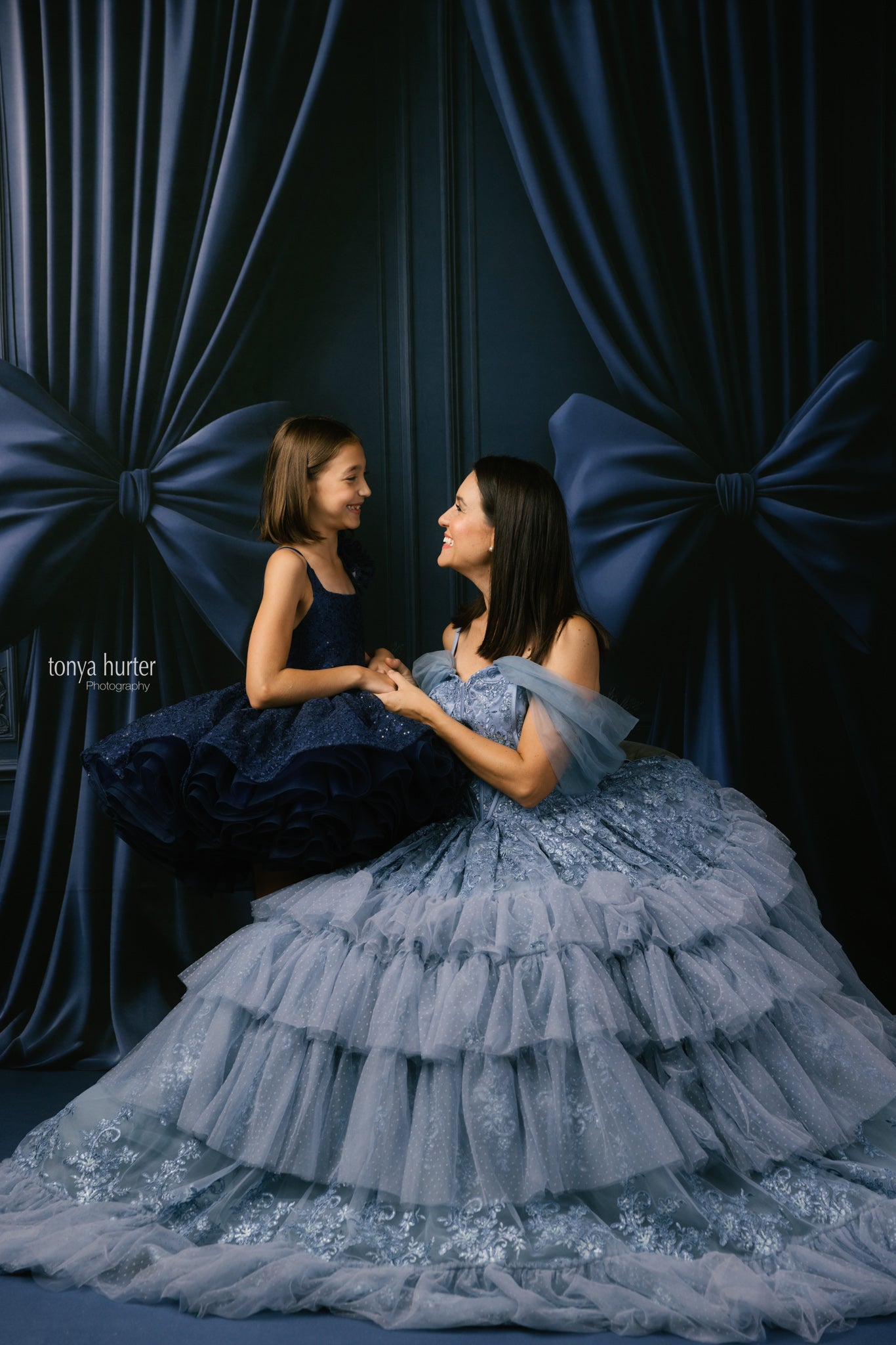 Two women in elegant dresses sitting against a dark curtain backdrop