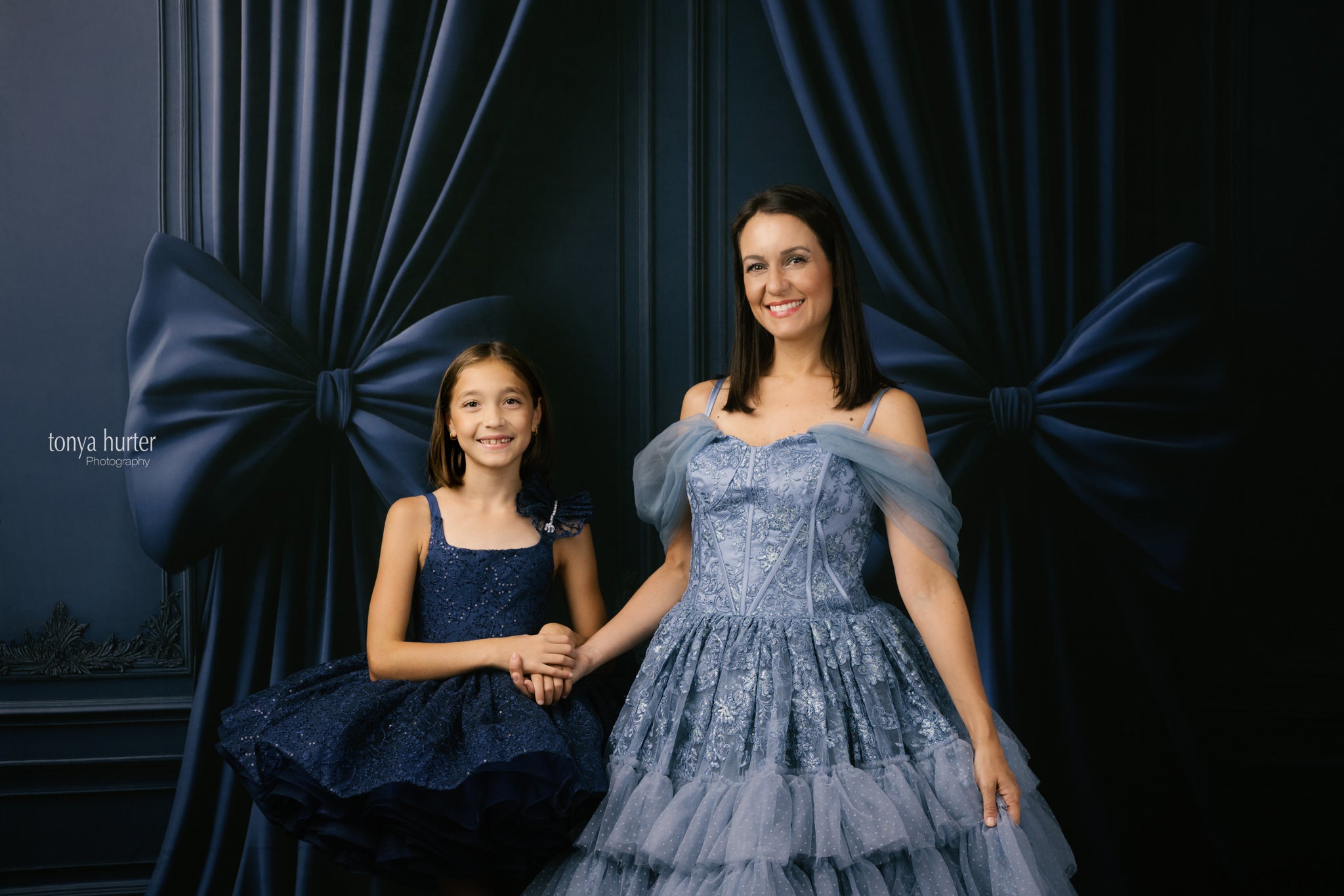 Two women in formal dresses standing in front of dark blue curtains with a bow.