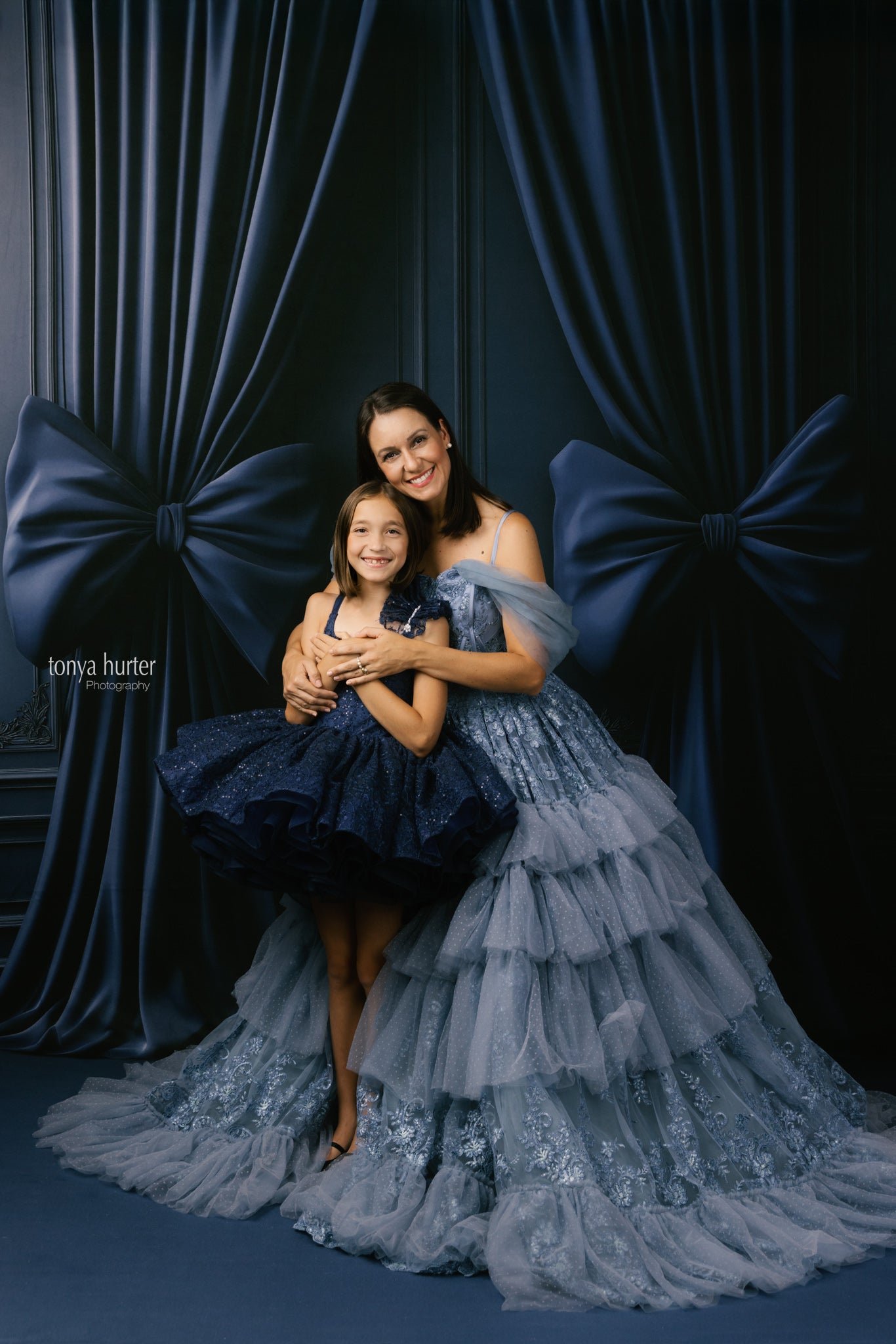 Two women in elegant dresses posing against a dark curtain backdrop