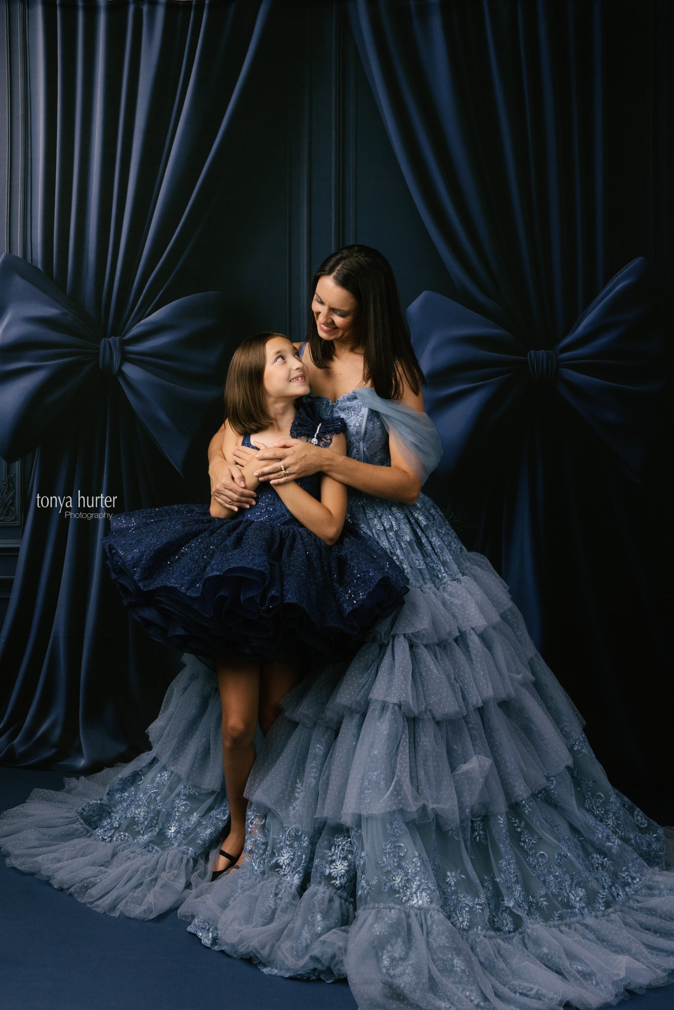 Two women in elegant dresses sitting together against a dark blue curtain backdrop.