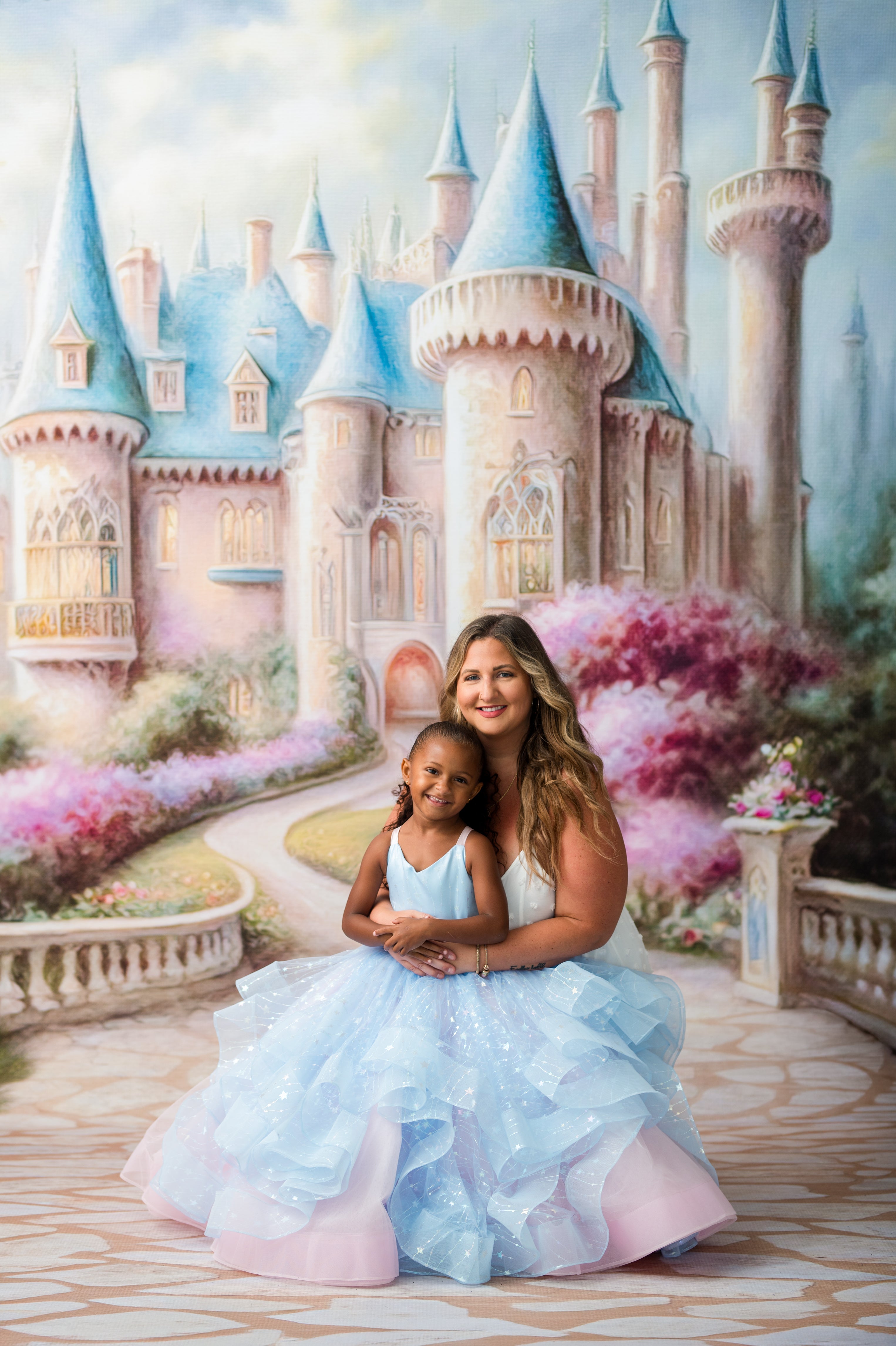 A woman and a young girl sitting together in front of a painted fairytale castle backdrop. The woman wears a voluminous light blue and pink gown and holds the child, who is dressed in a matching light blue gown.