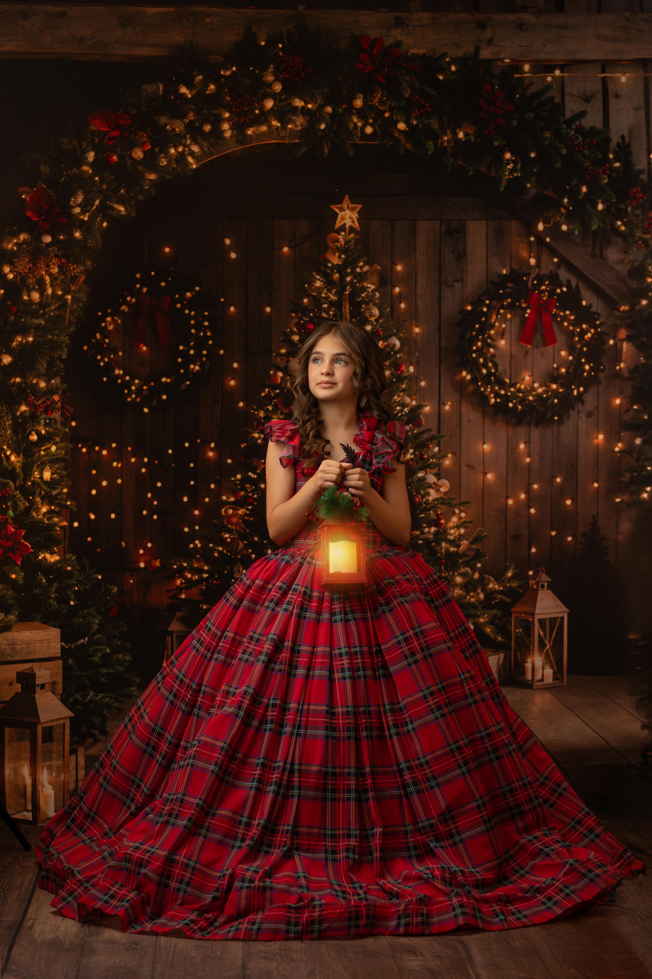 Child wearing a festive plaid holiday dress, holding out the skirt, with a Christmas tree decorated with lights in the background.