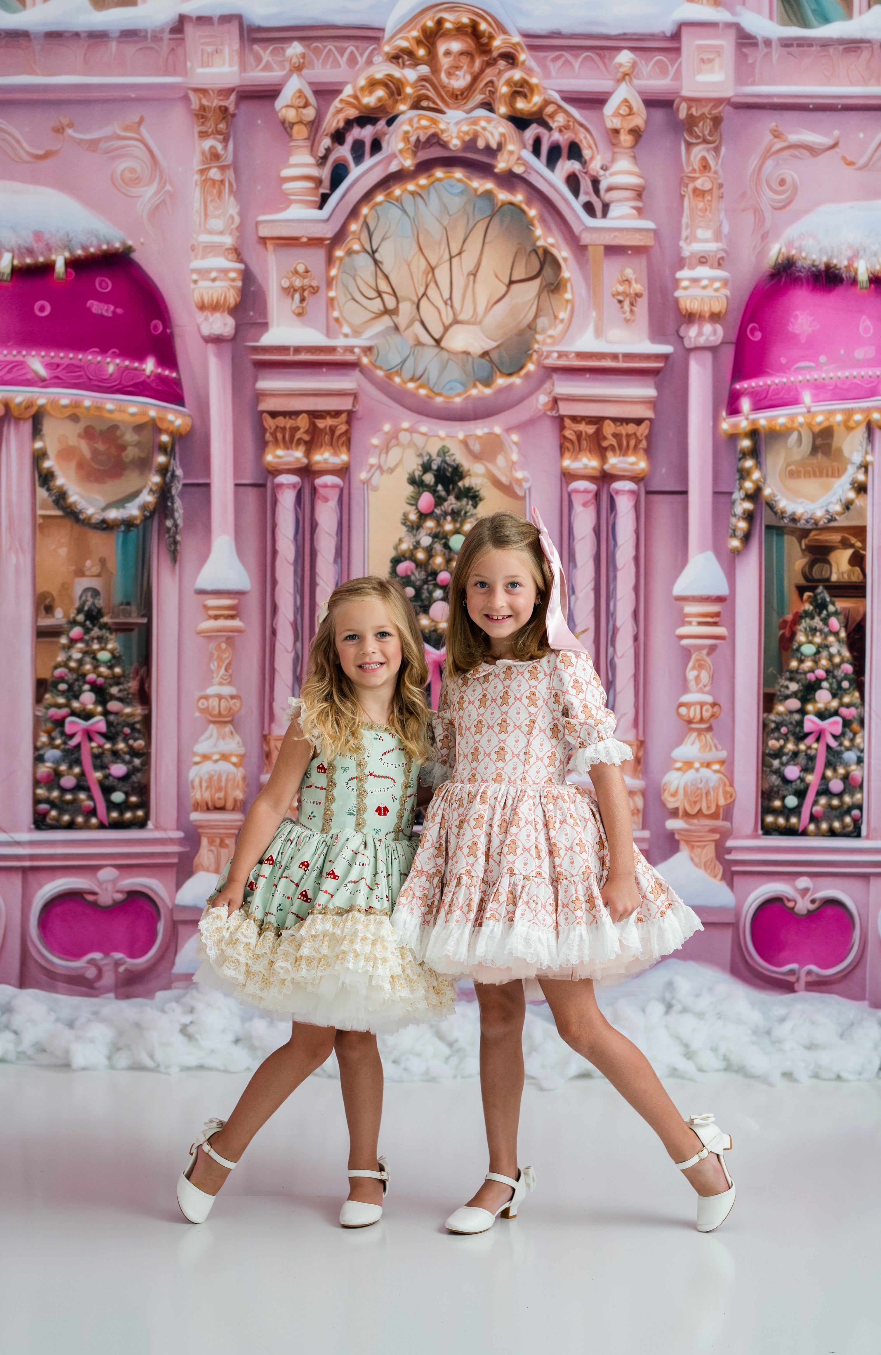 Two young girls posing in front of a whimsical, pink and gold Christmas-themed backdrop that resembles an ornate candy shop facade. The girl on the left wears a light green and white patterned dress with white tulle, while the girl on the right wears a peach and white patterned dress with white tulle.
