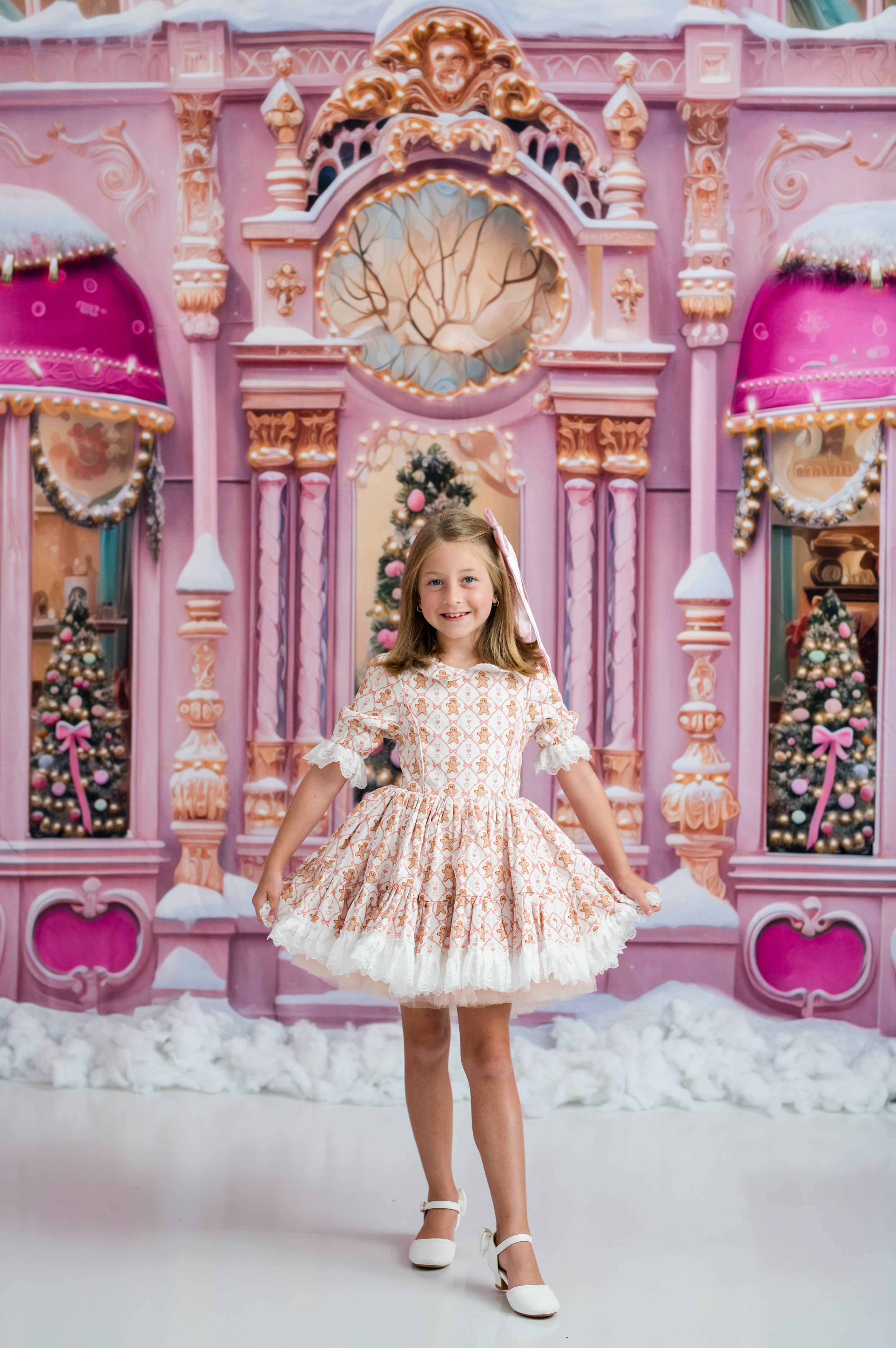 A young girl stands in front of a large pink holiday-themed backdrop, wearing a short cream dress with pink floral patterns and a ruffled white hem, paired with white shoes.
