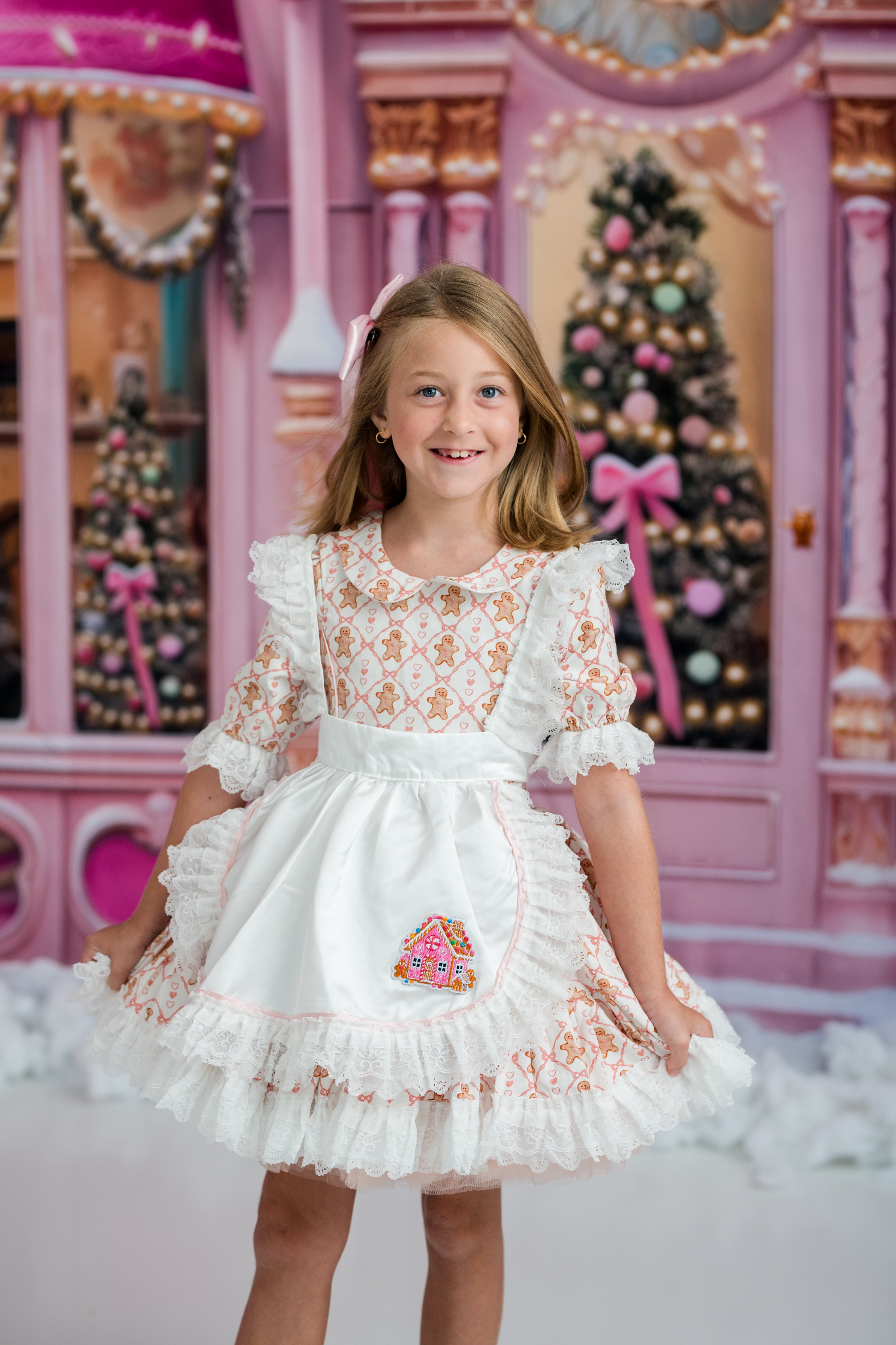 A young girl with blonde hair wearing a white and pink ruffled dress with a gingerbread-themed bodice and apron pocket, standing in front of a pink holiday-themed backdrop with Christmas trees.
