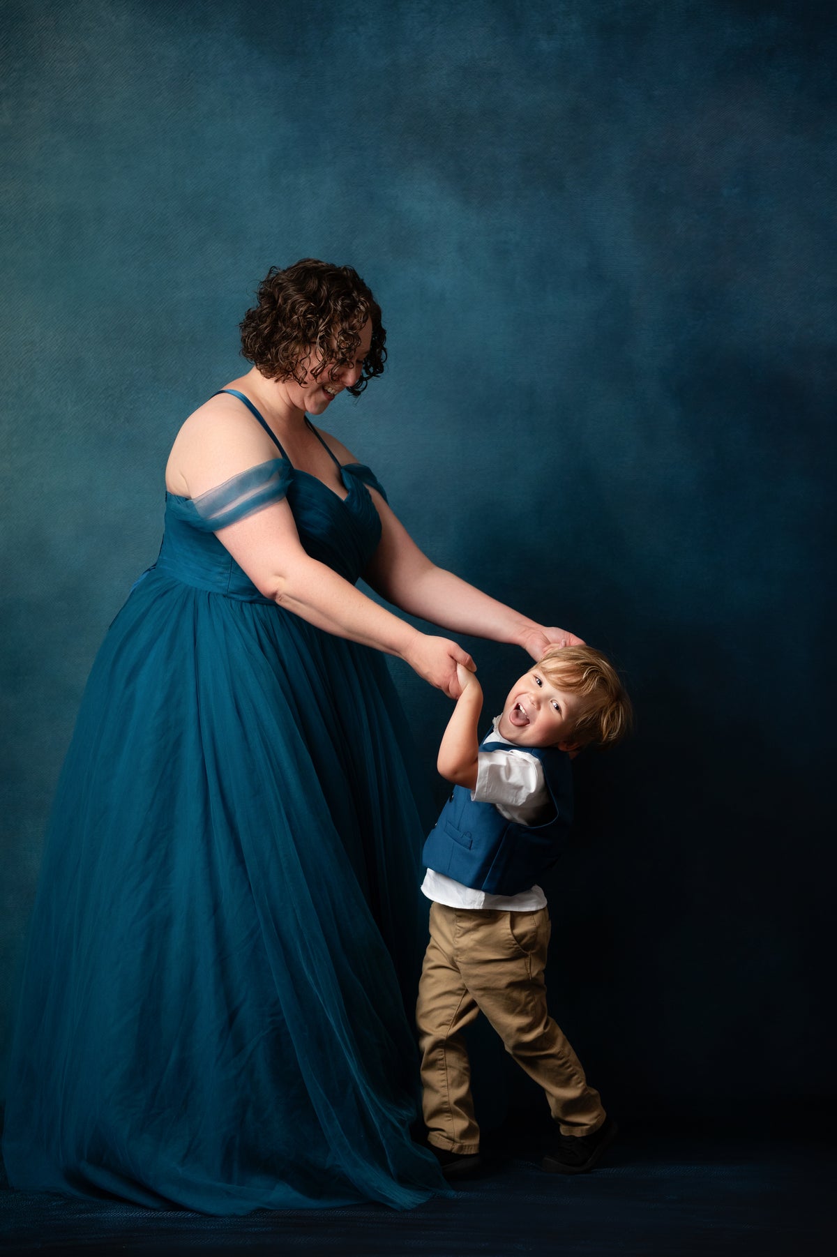 A woman in a floor-length teal blue gown holds a young boy who is wearing a blue vest, white shirt, and tan pants, posed against a dark blue studio backdrop.