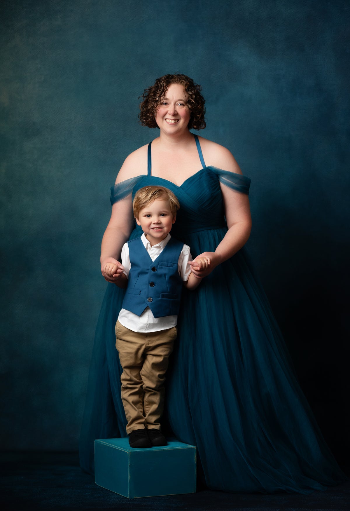 A woman in a floor-length teal blue gown stands behind a young boy wearing a blue vest, white shirt, and tan pants, holding his hands as he stands on a matching teal block against a dark blue studio backdrop.