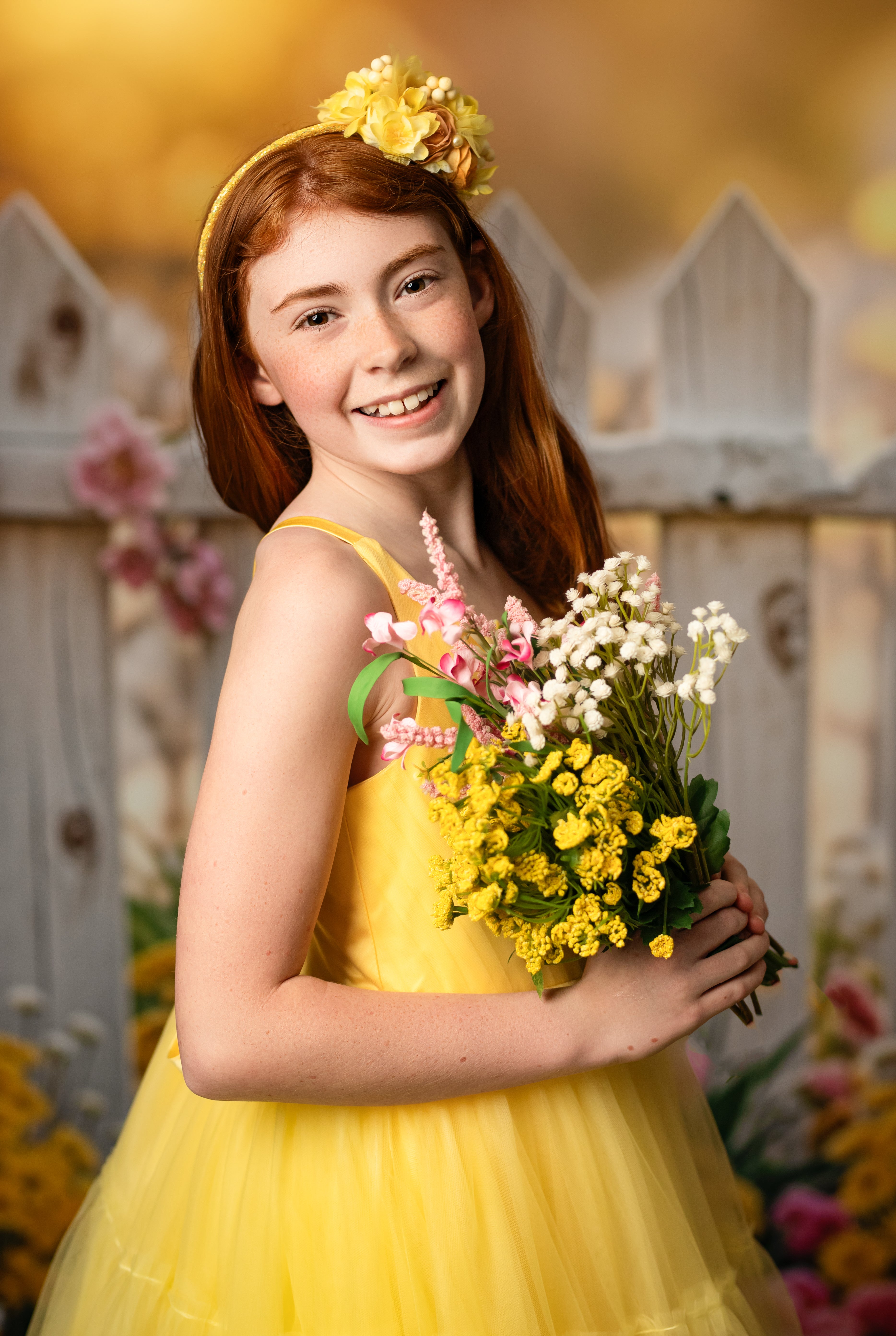 Golden glow – The warmth of the golden hour enhances the beauty of a girl in a tulle gown, standing softly against a floral-accented fence.