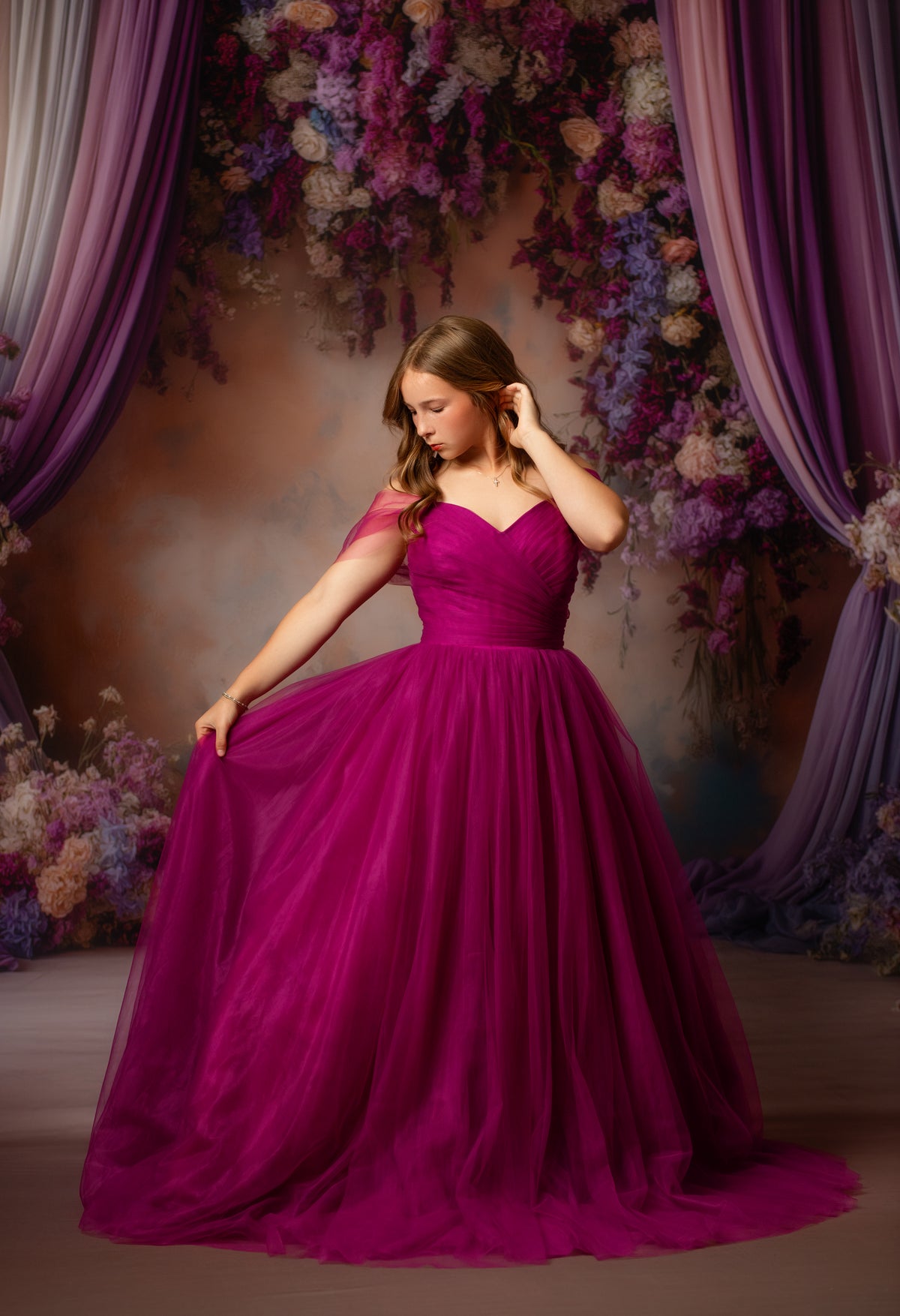 A young woman wearing a voluminous magenta ball gown with sheer cap sleeves, posing in front of a purple floral backdrop with draped curtains.