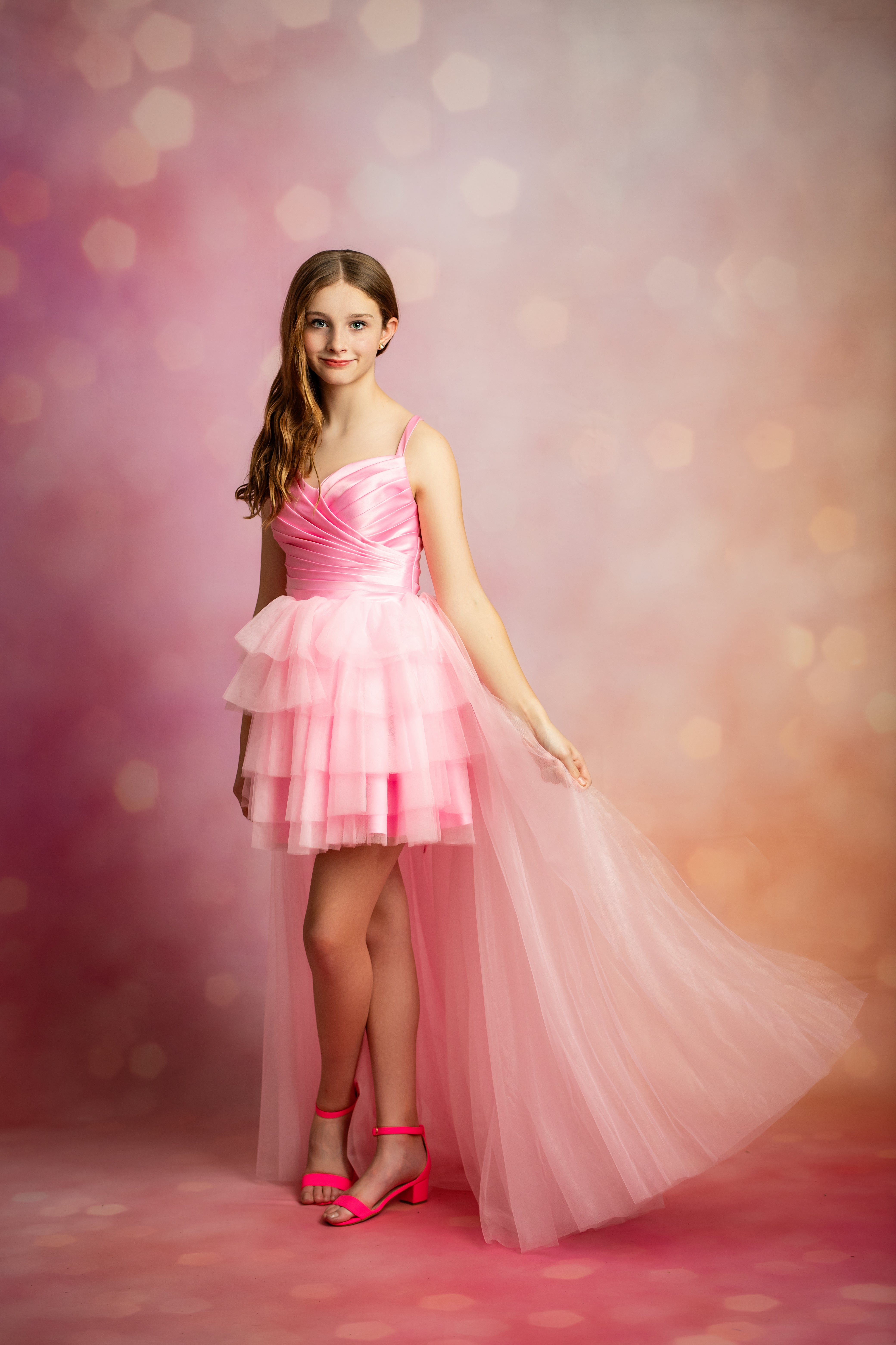 A young girl models a pink strapless tulle dress with a ruffled tiered skirt and a long flowing train, standing against a pink and orange bokeh studio backdrop.