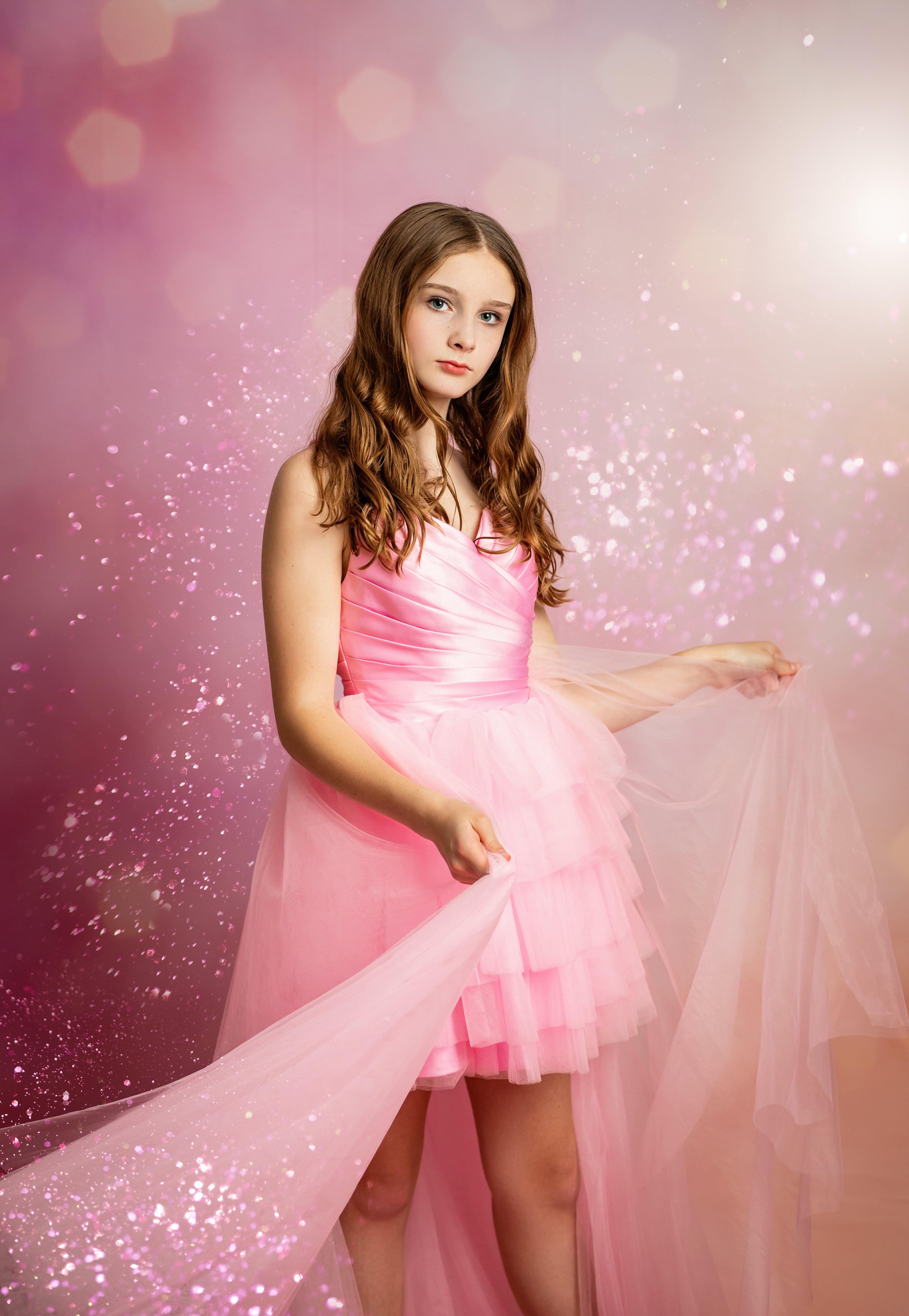 A young girl with long brown hair wearing a pink strapless tulle dress, holding out a sheer pink fabric against a sparkling pink and purple studio background.