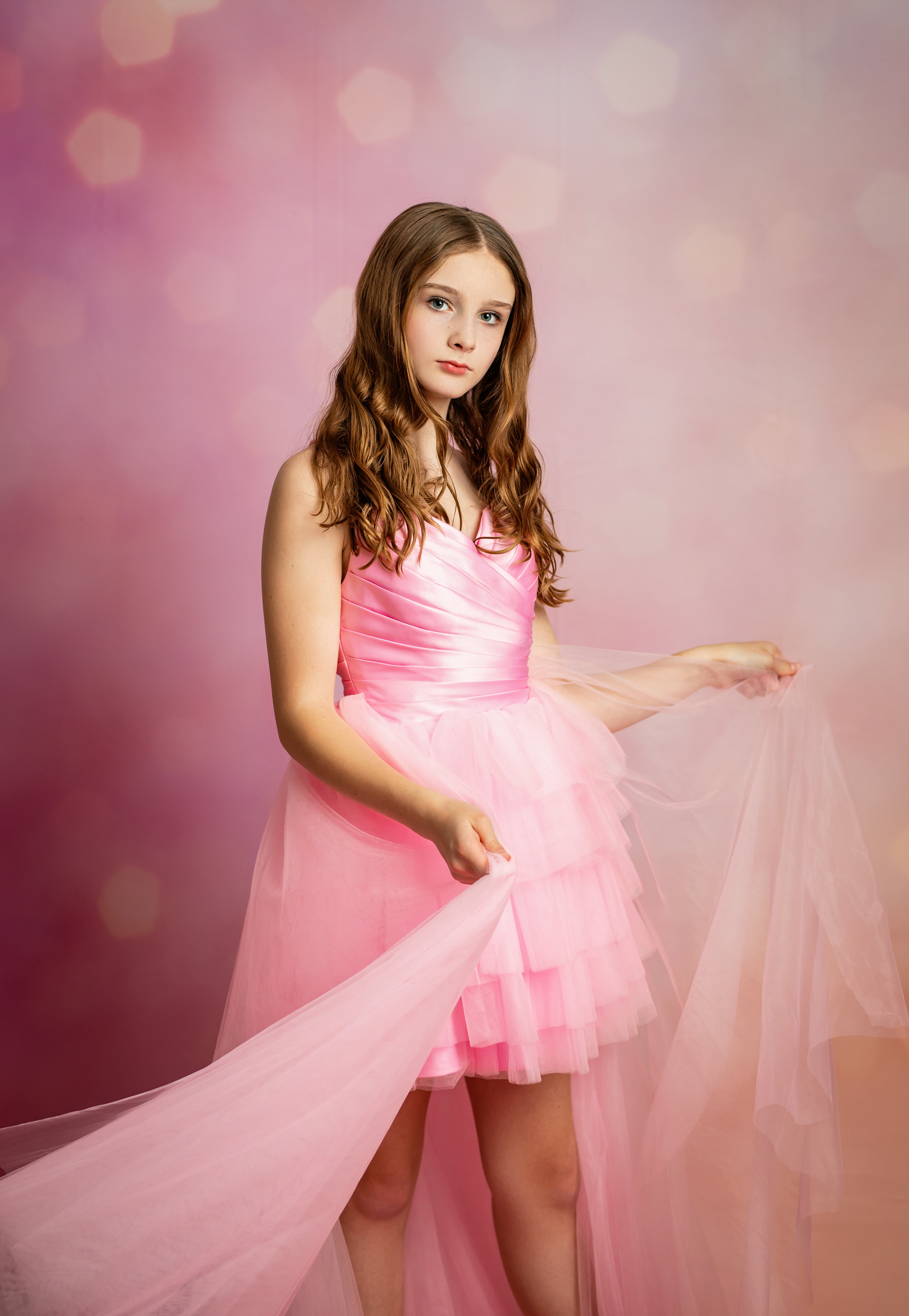 A young girl with long brown hair wearing a pink satin-strap dress with a layered tulle skirt, holding out a sheer pink fabric against a soft pink bokeh studio background.