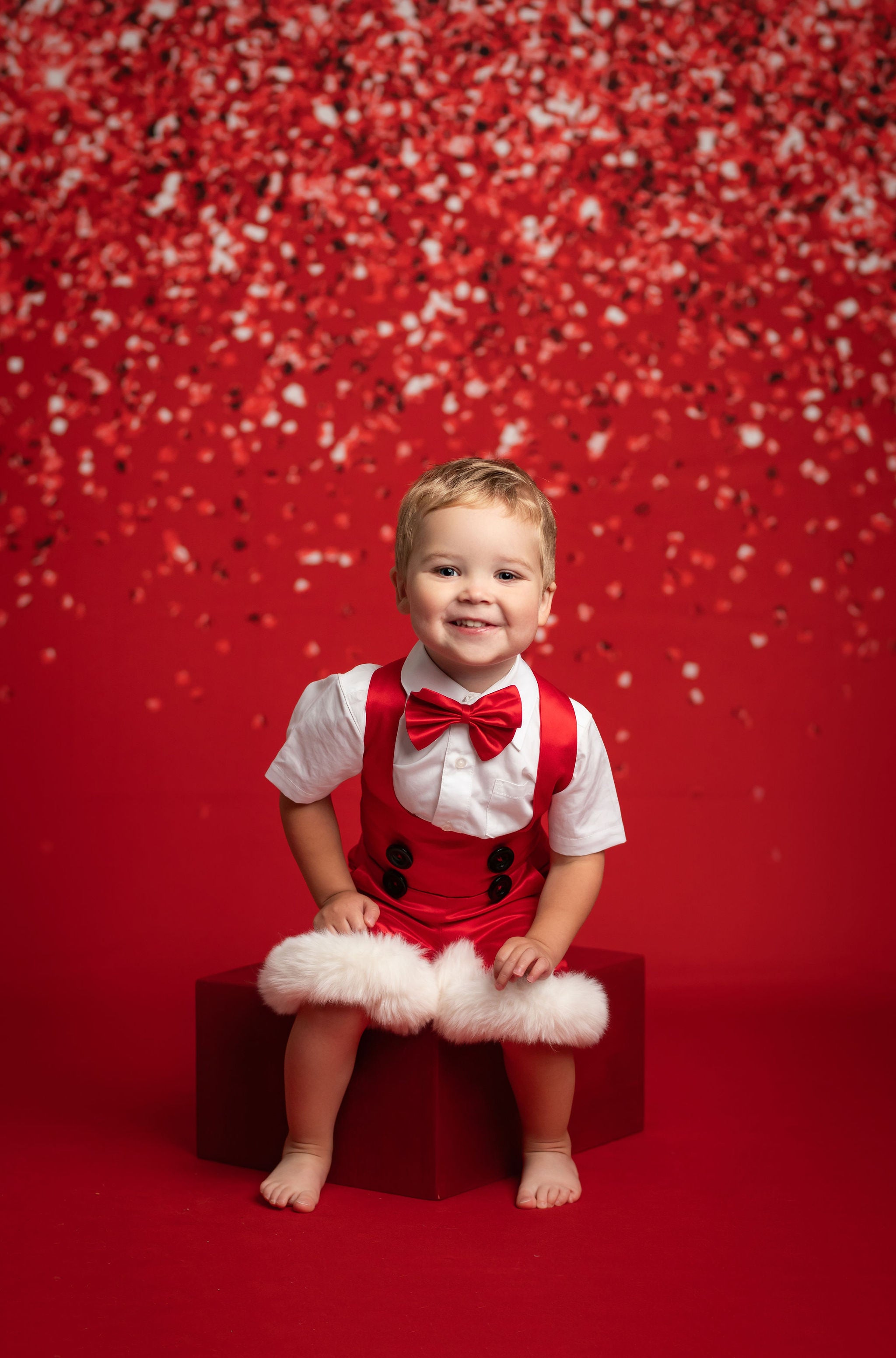 Child in a red outfit with a bow tie sitting on a red box against a red background with confetti.
