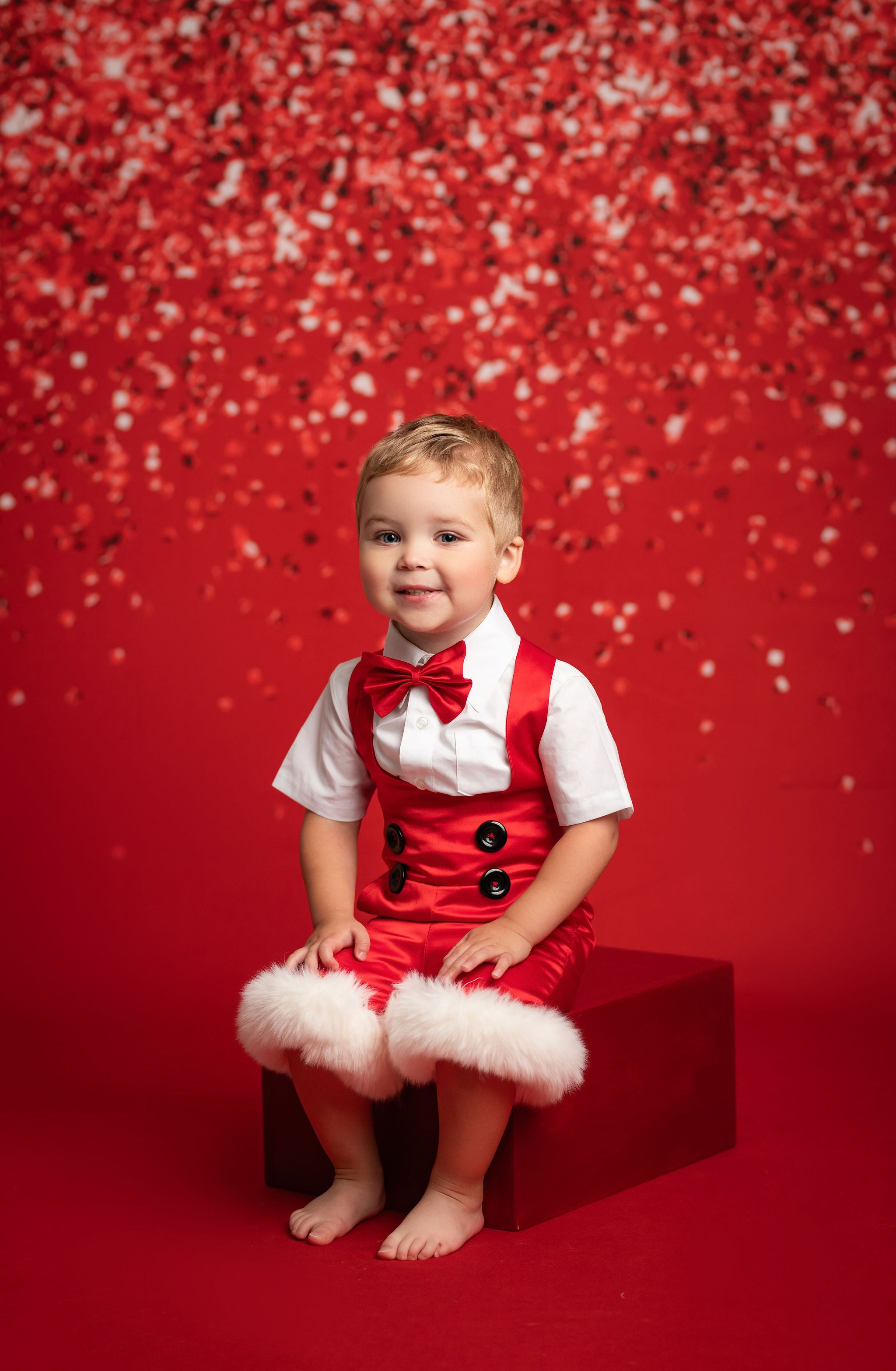 Child in a red outfit with a bow tie sitting on a red box against a red background with confetti.