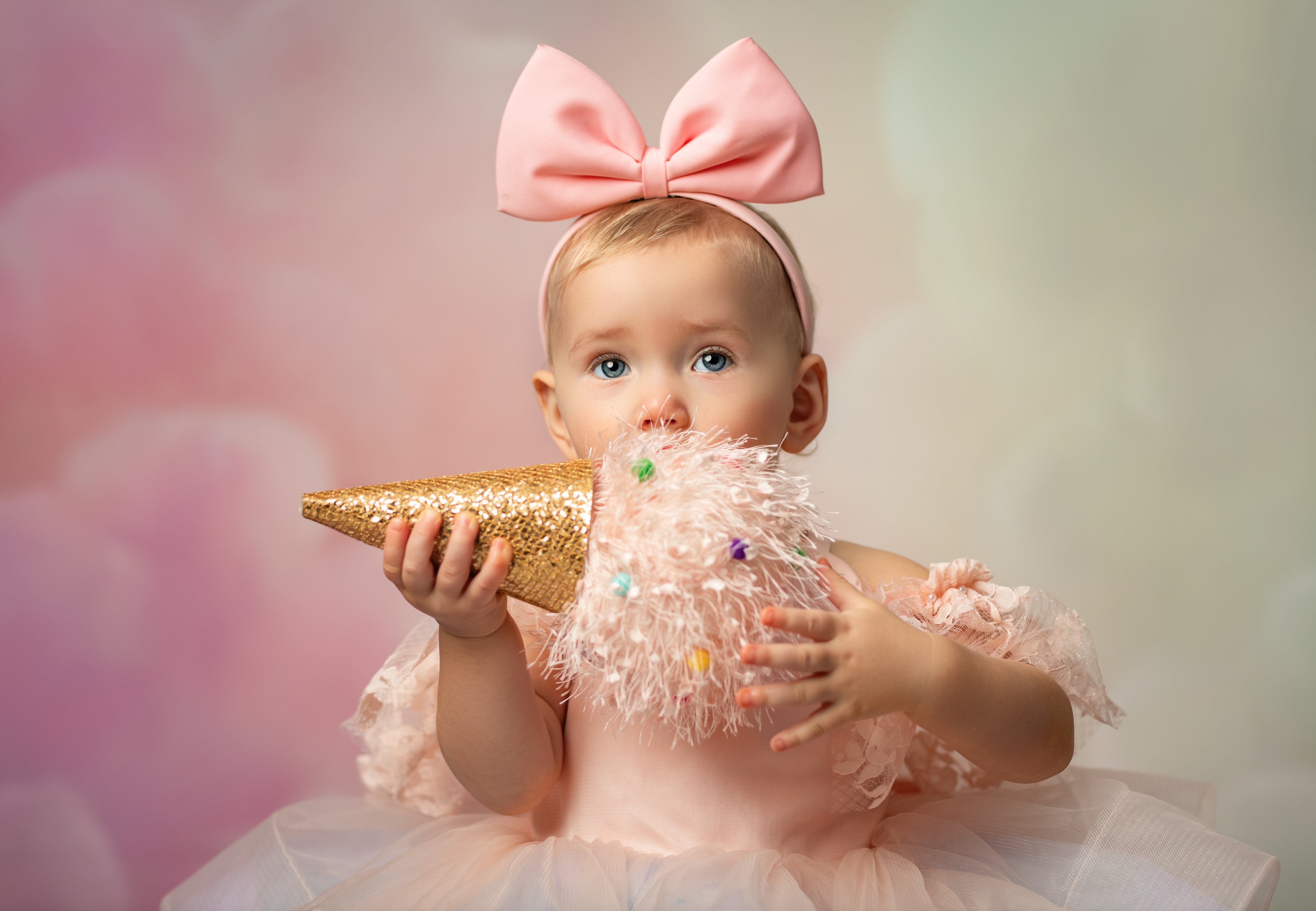 Baby in a pink dress with a large bow holding a gold glittery cone.