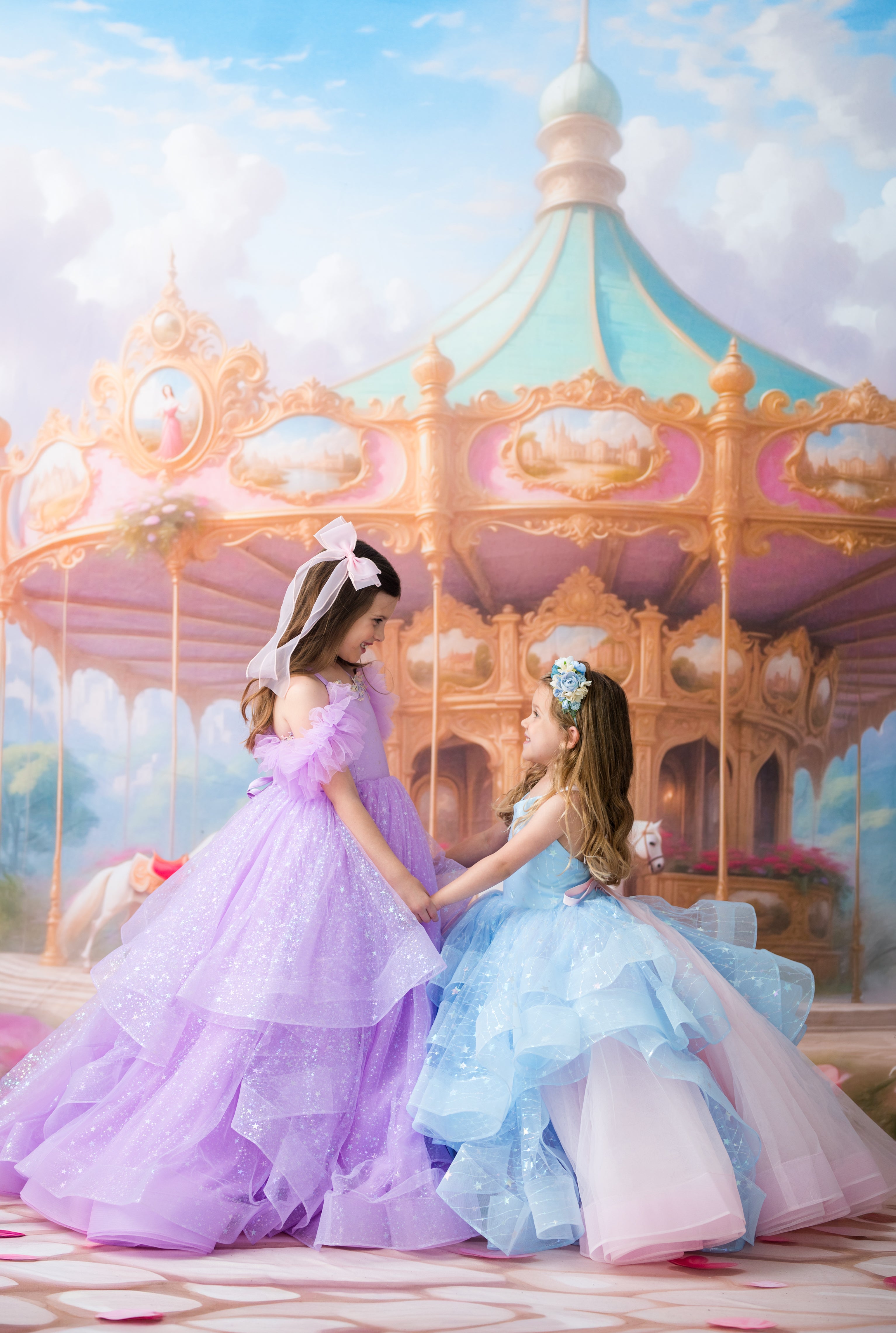 Two young girls in elegant, sparkling ballgowns pose together in front of a whimsical pastel carousel. The girl on the left wears a lavender-purple gown with a white bow, and the girl on the right wears a light blue gown with a pink underskirt.