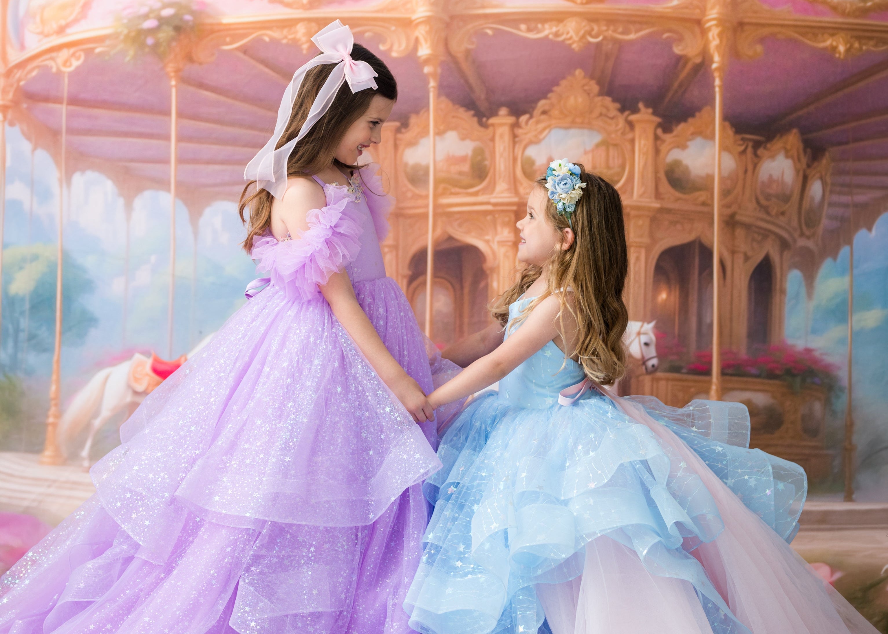 Two young girls in elegant, sparkling ballgowns pose together in front of a whimsical pastel carousel. The girl on the left wears a lavender-purple gown with a white bow, and the girl on the right wears a light blue gown with a pink underskirt.