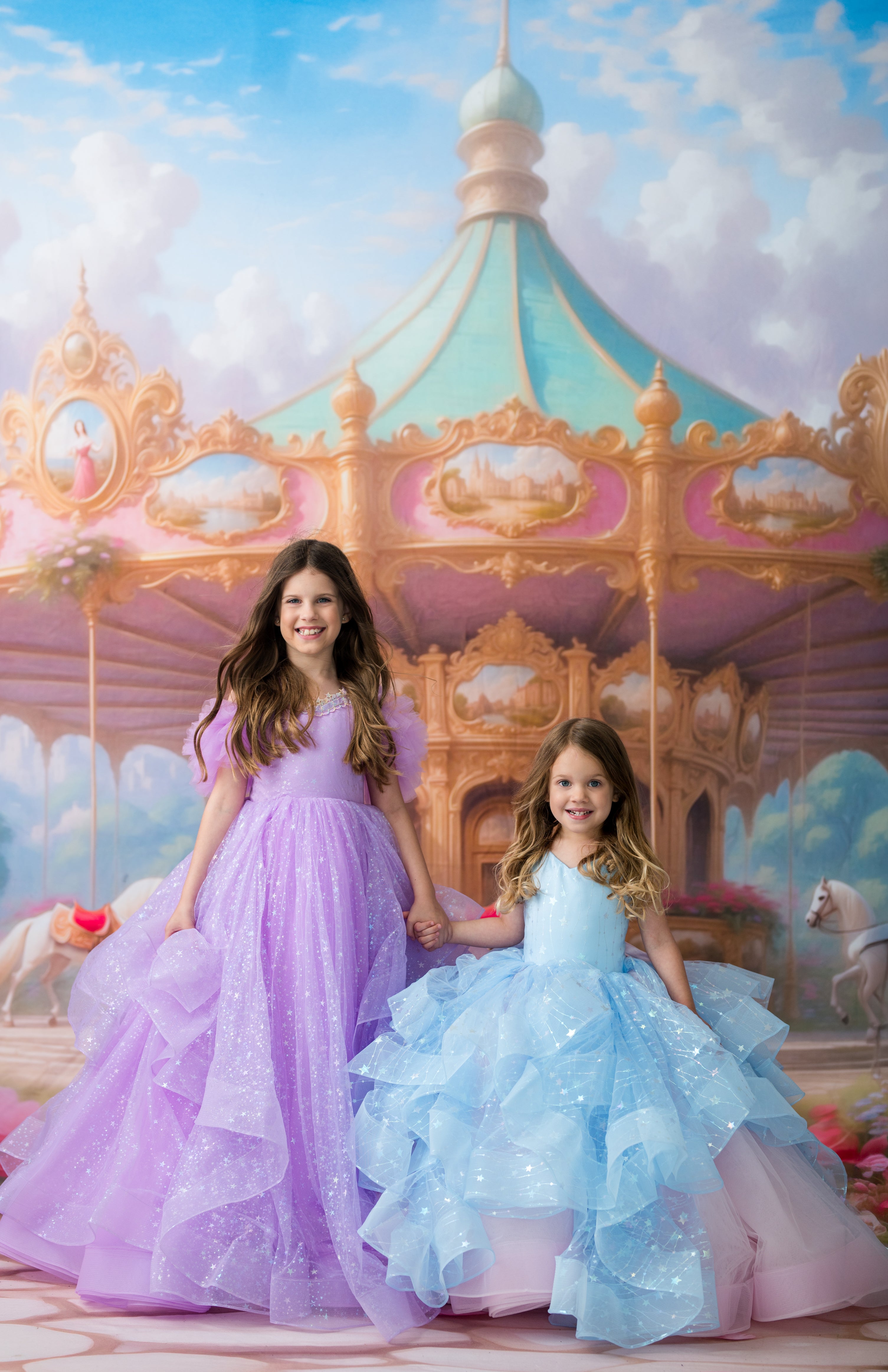 Two young girls in princess gowns posing in front of a painted carousel backdrop. The girl on the left wears a lavender-purple gown, and the girl on the right wears a light blue ruffled gown with pink underlayer.