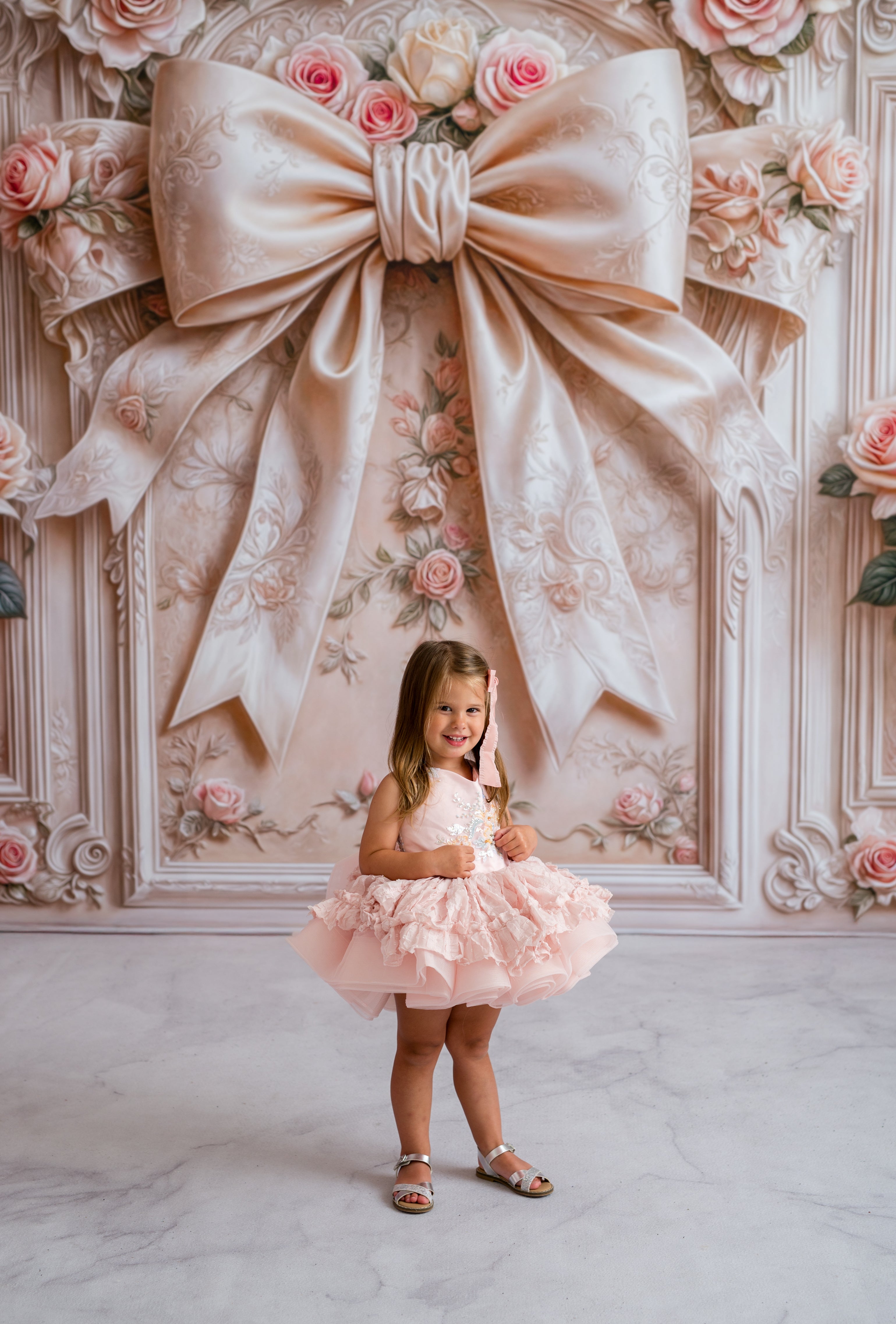 Young girl in a pink dress standing in front of a decorative wall with a large bow and flowers.