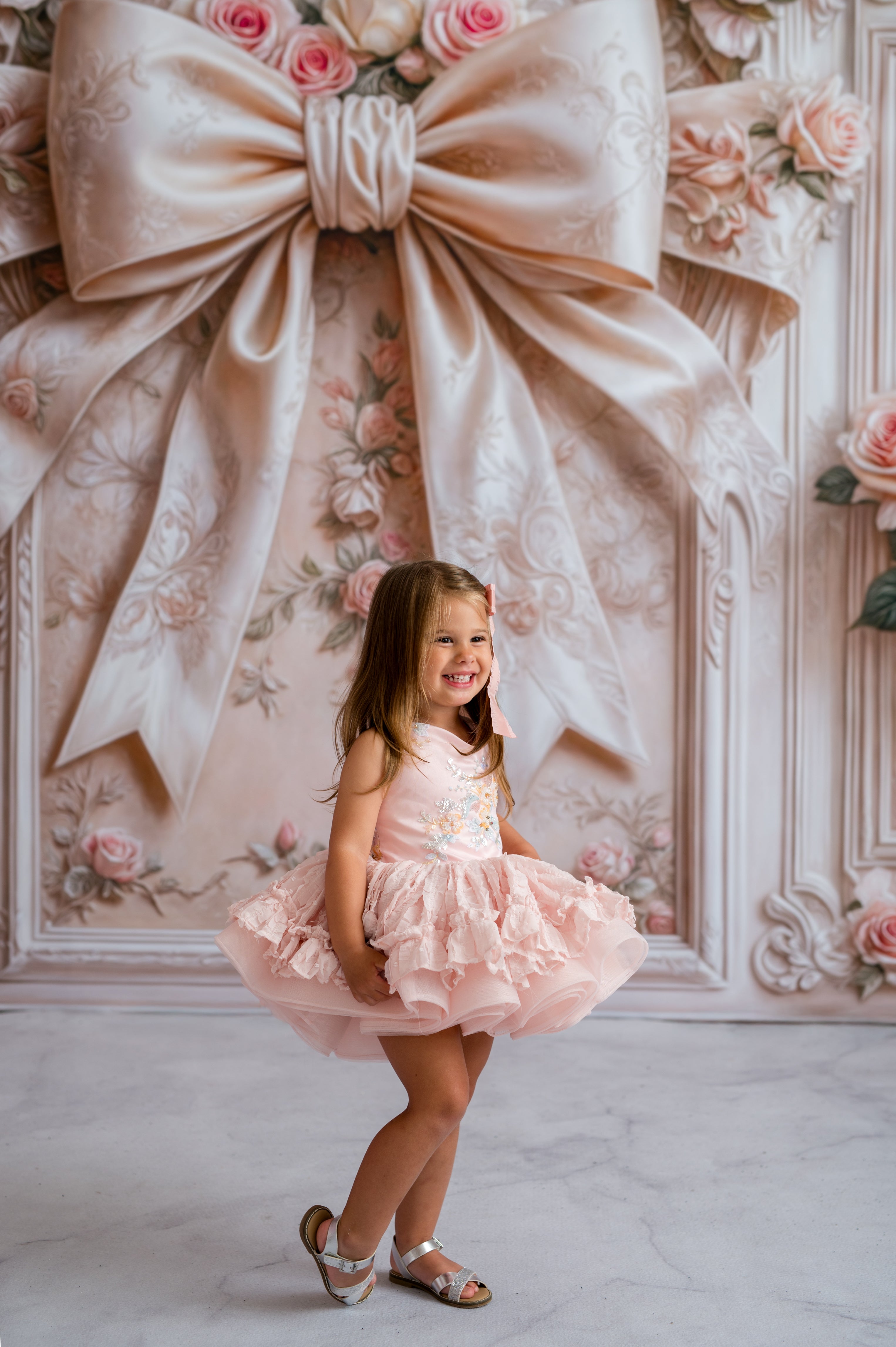 Young girl in a pink dress standing in front of a decorative wall with a large bow and floral patterns.
