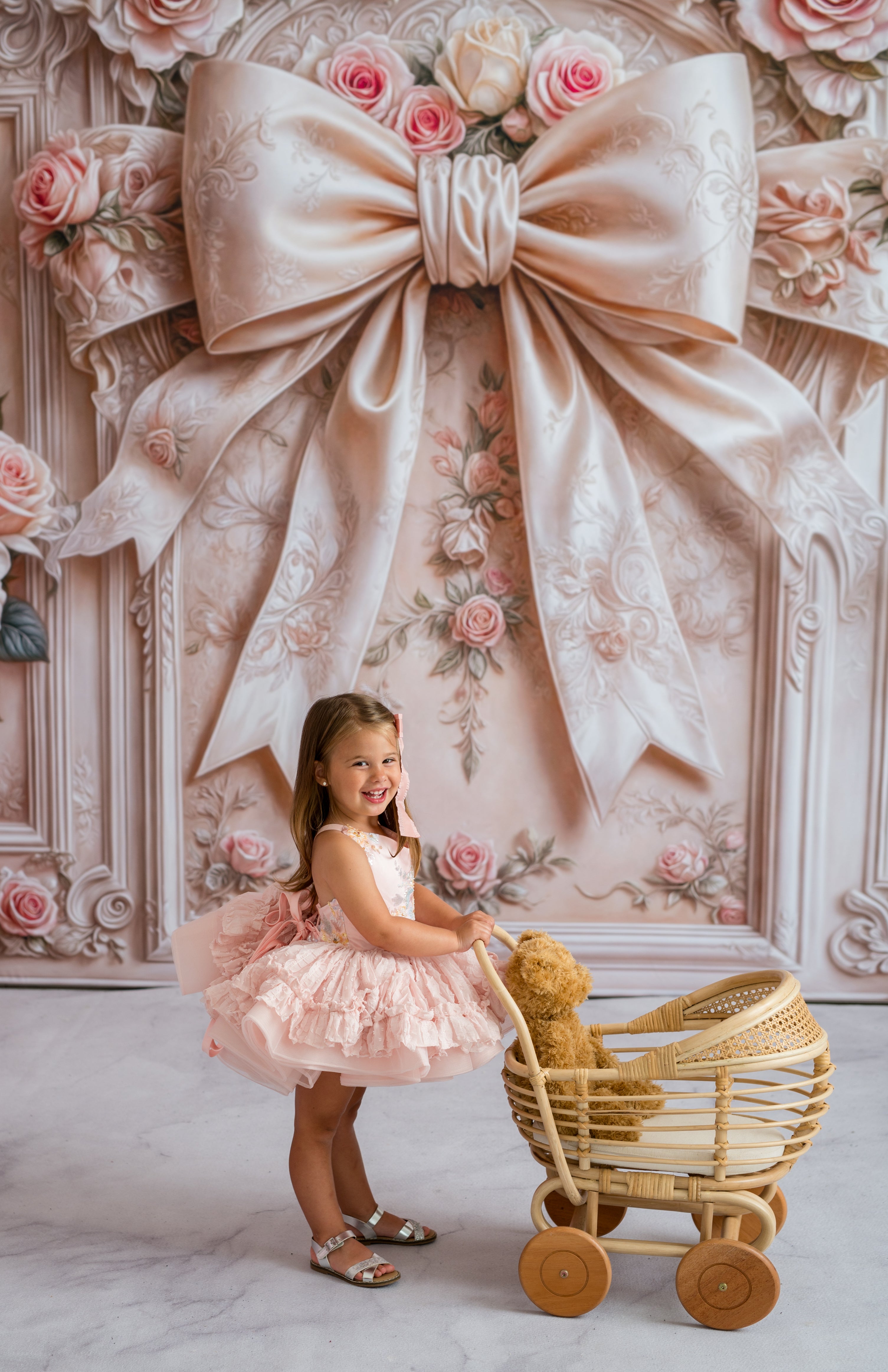 Young girl in a pink dress standing next to a wooden pram in front of a decorative wall with a large bow and flowers.