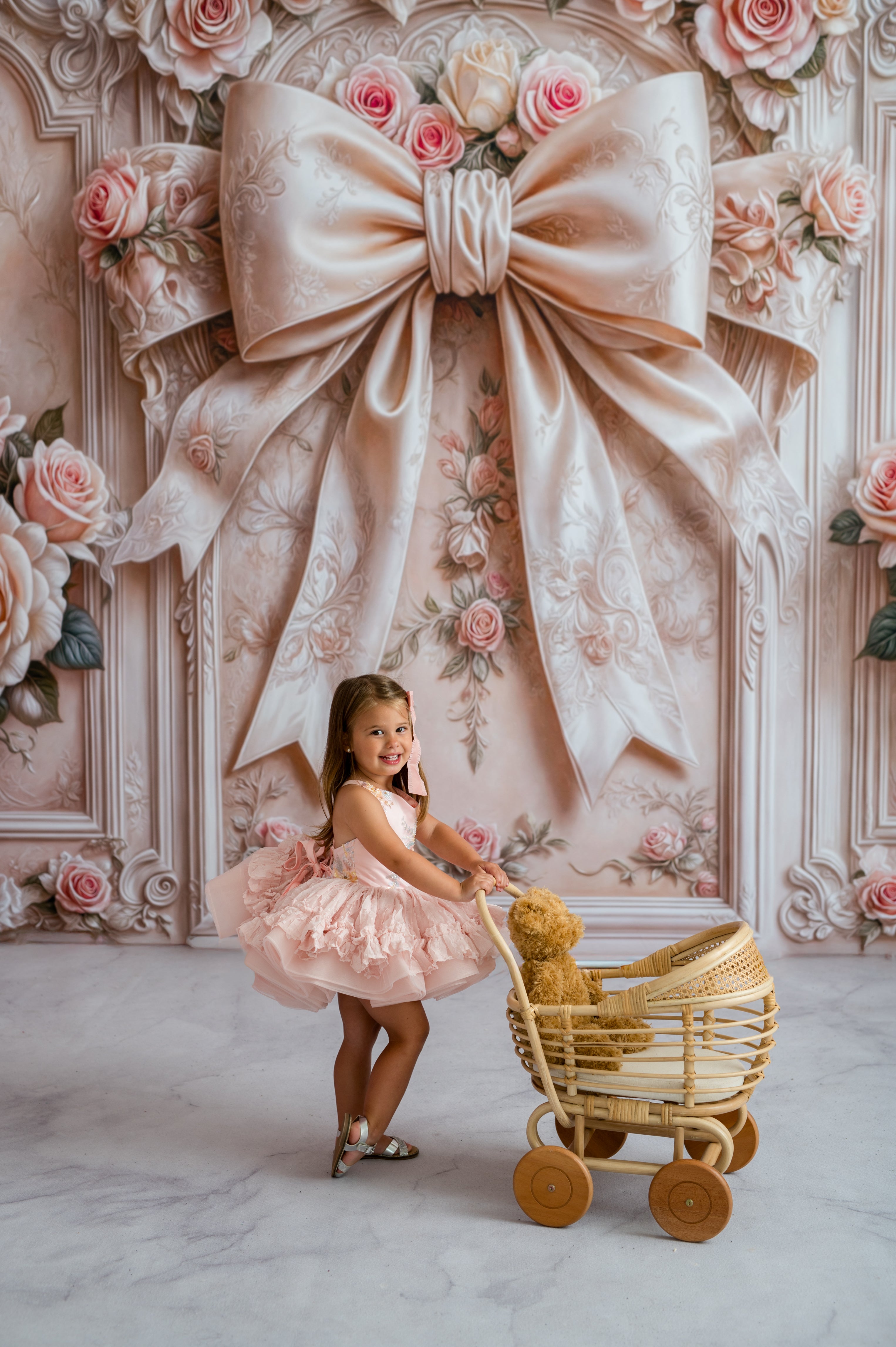 Young girl in a pink dress pushing a toy stroller with a large decorative bow and flowers in the background.
