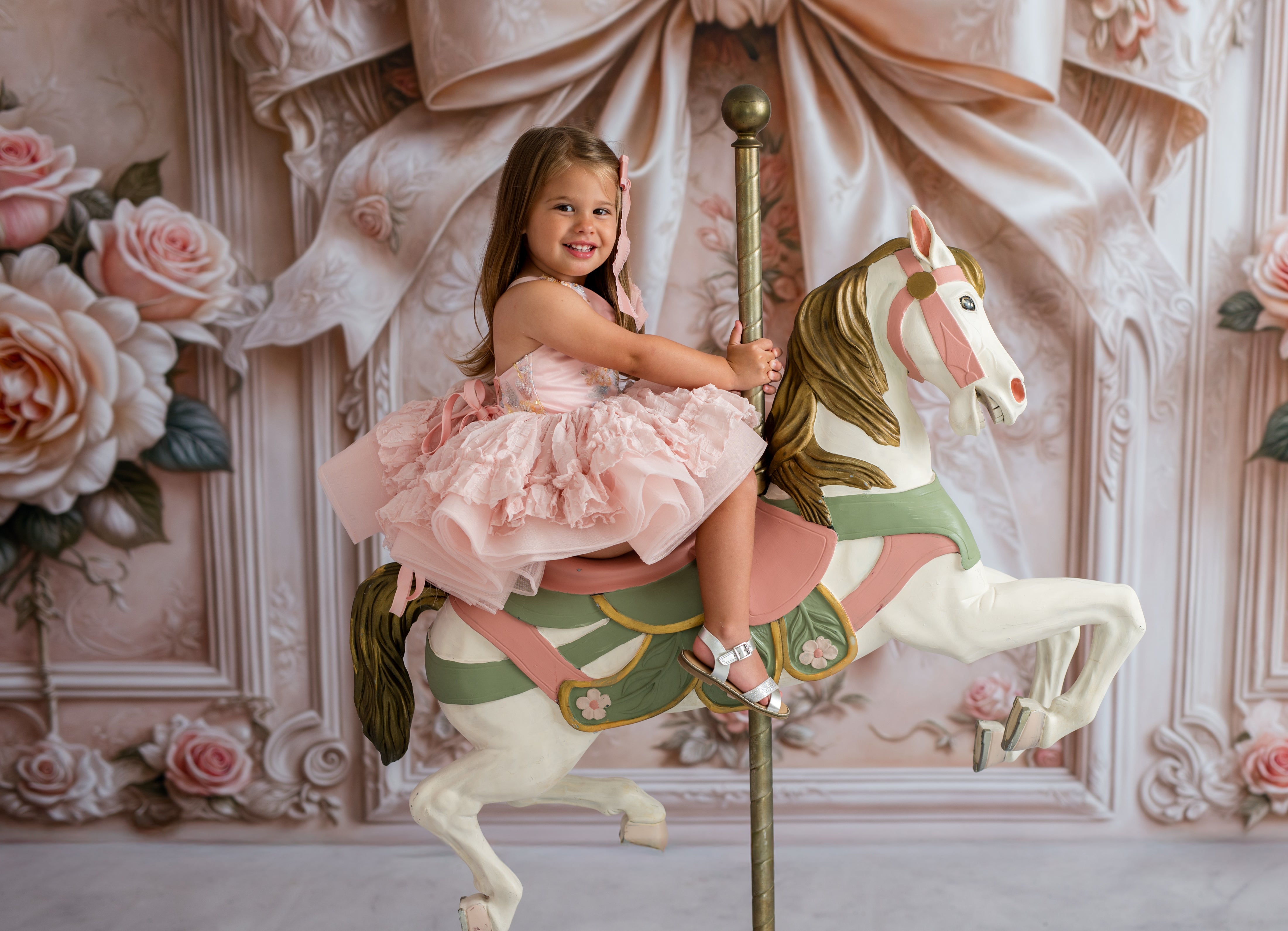 Young girl in a pink dress riding a carousel horse against an ornate wall with floral decorations.