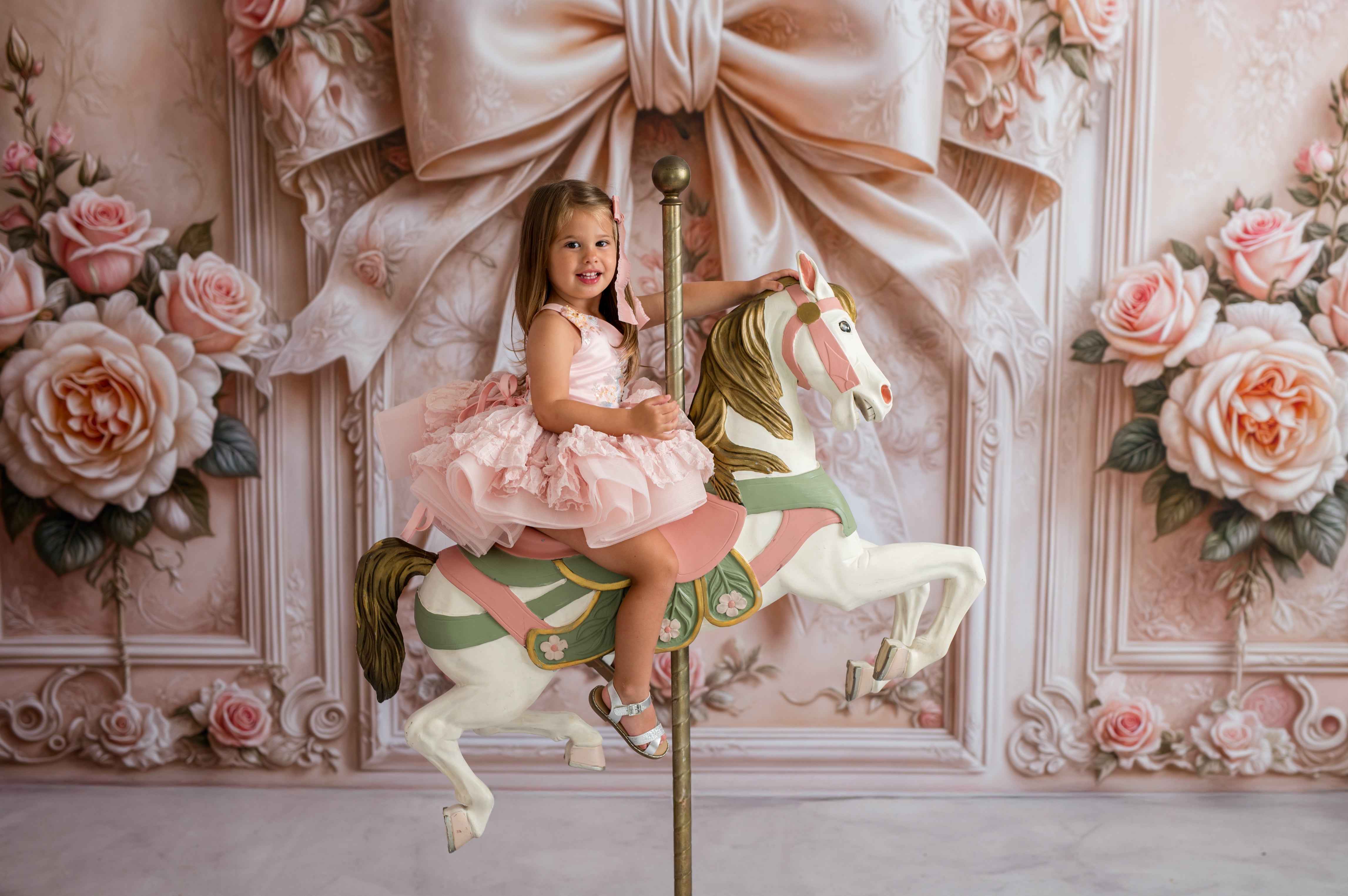 Young girl in a pink dress riding a carousel horse against a decorative floral wall.