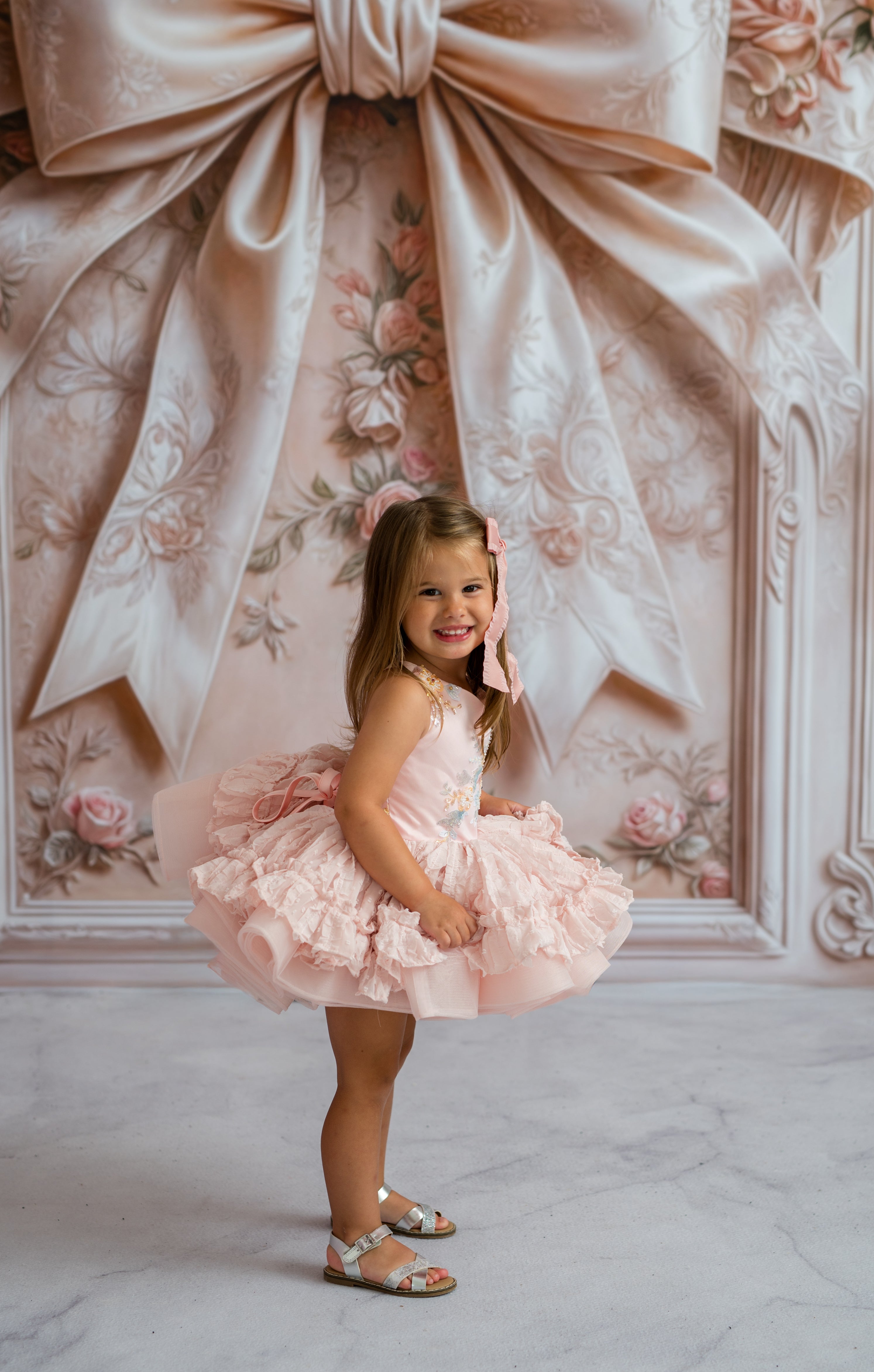 Young girl in a pink dress standing in front of a decorative wall with a large bow.