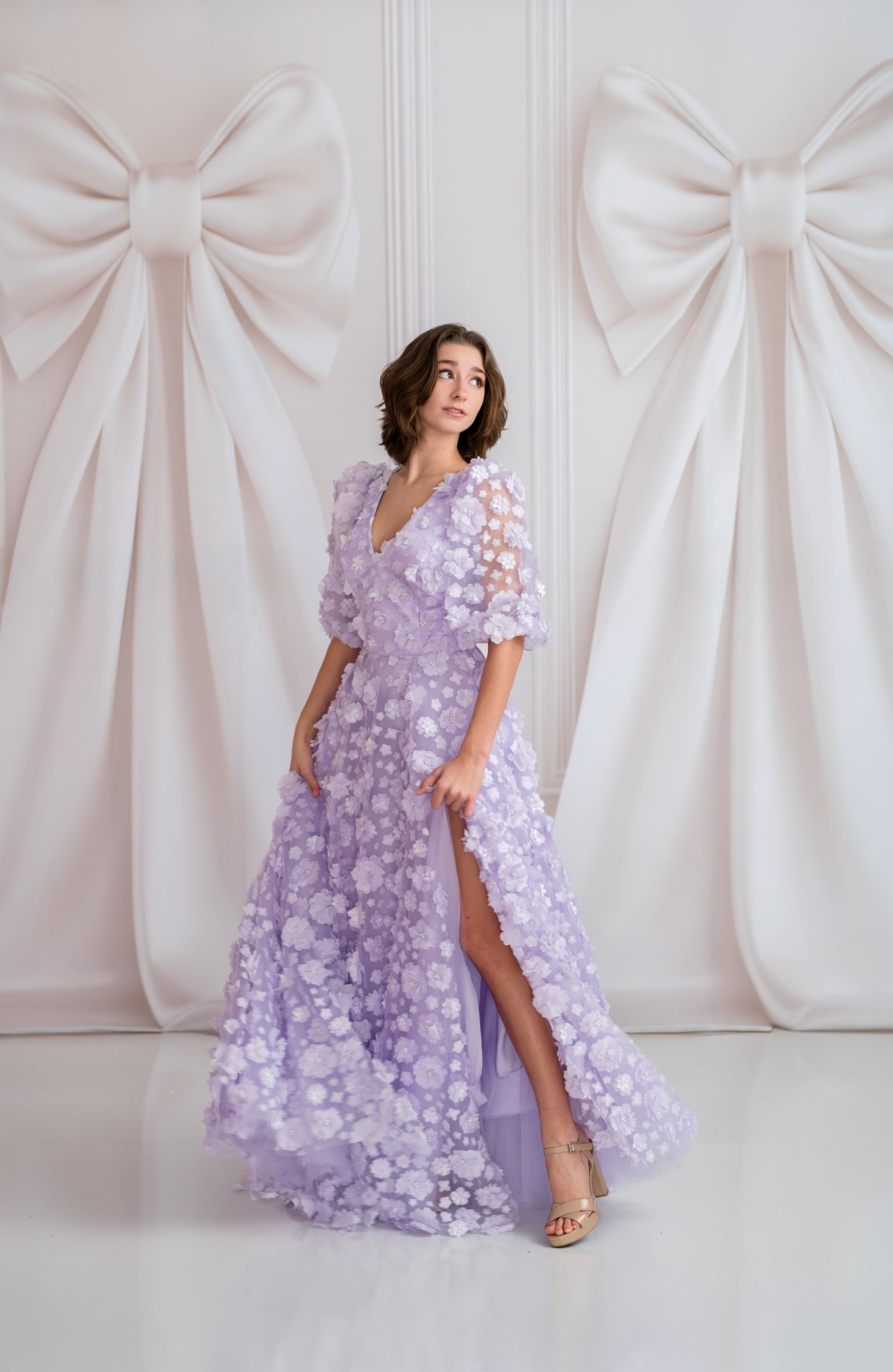 Woman in a lavender floral dress standing against a decorative white wall with large bows.