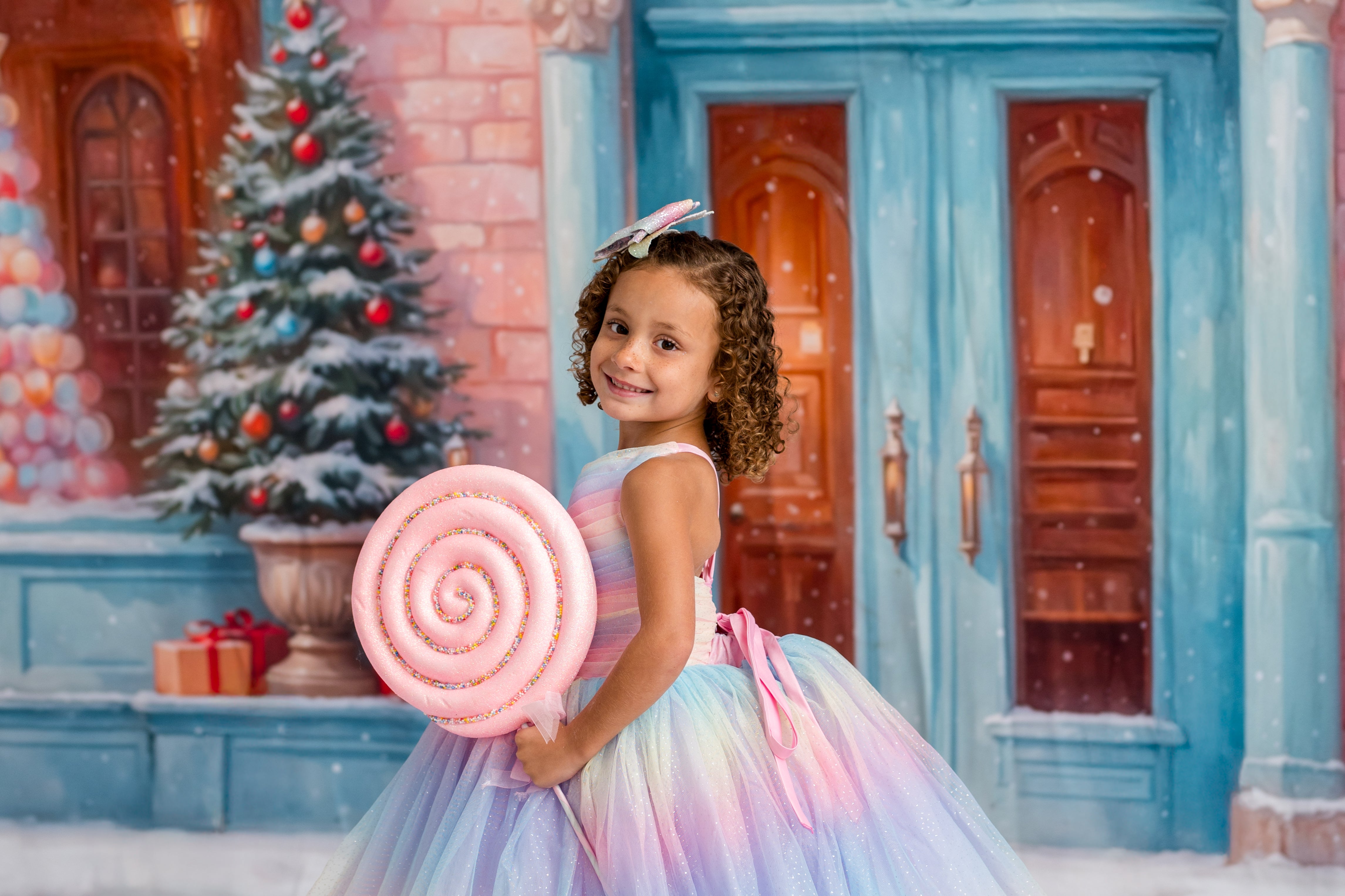 Young girl in a colorful dress holding a large candy cane in a festive setting with a Christmas tree and presents.