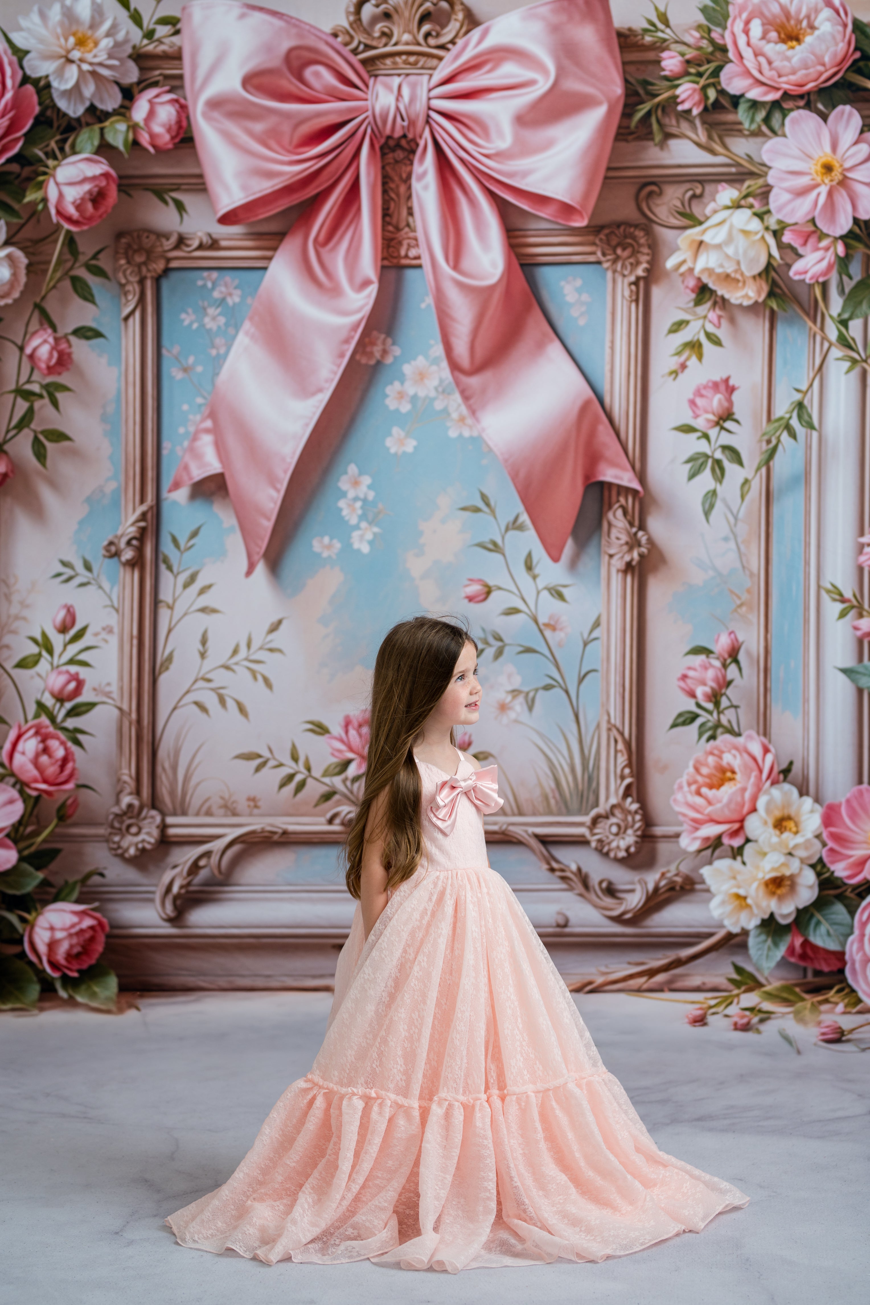 Young girl in a pink dress standing in front of a decorative wall with flowers and a large bow.